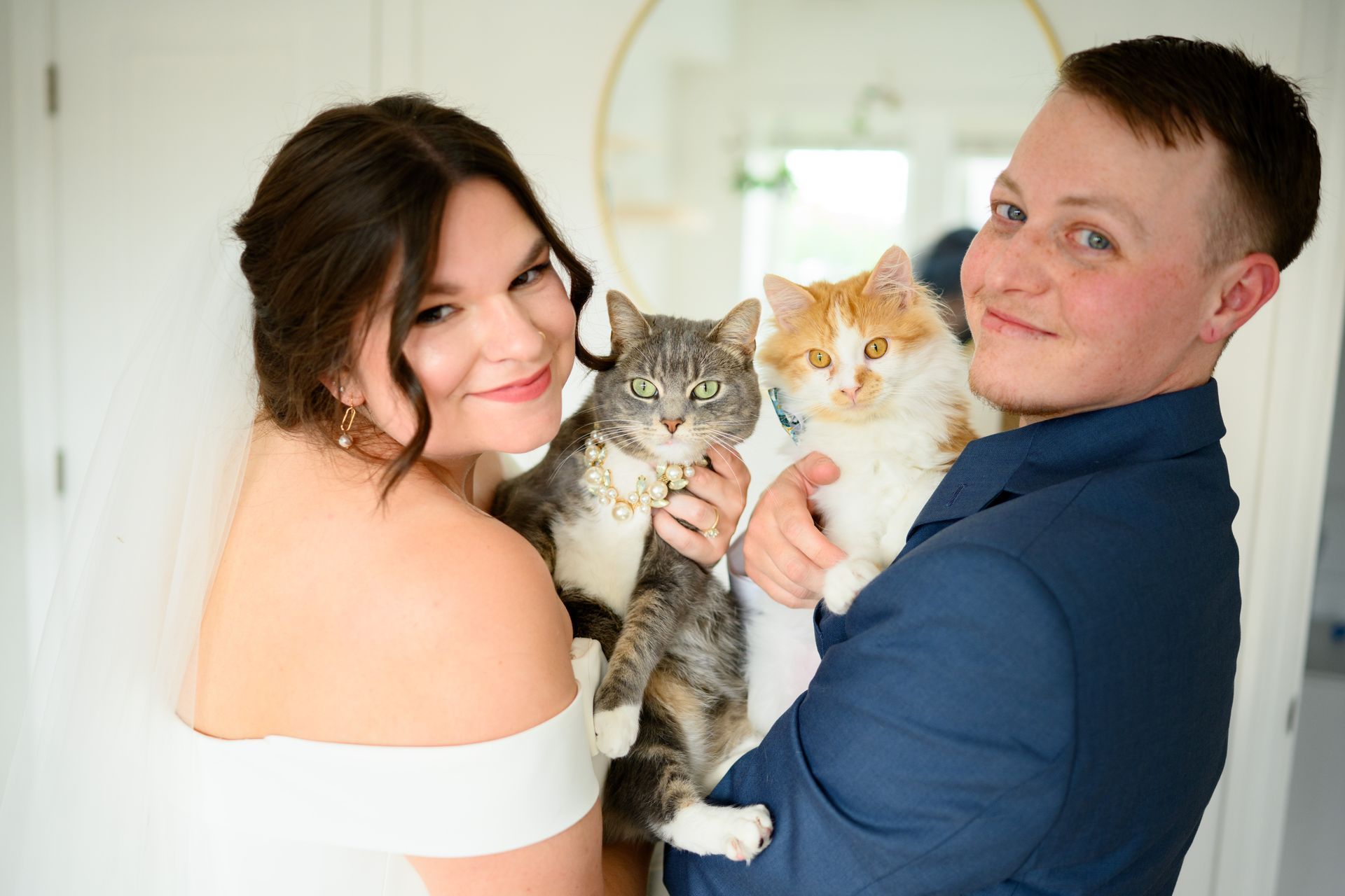 Bride and groom holding two cats; bride in white dress, groom in blue suit, both smiling.