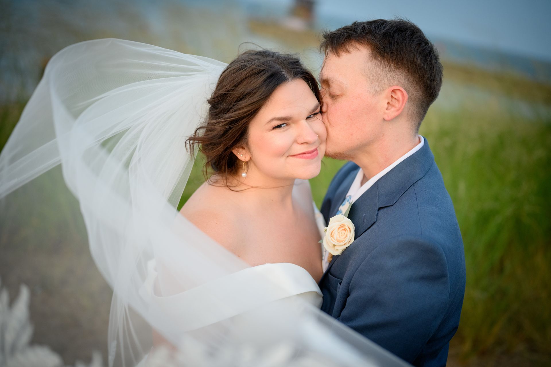 Bride and groom embrace, he kisses her cheek. She wears a veil and white dress, he wears a blue suit; outdoors.