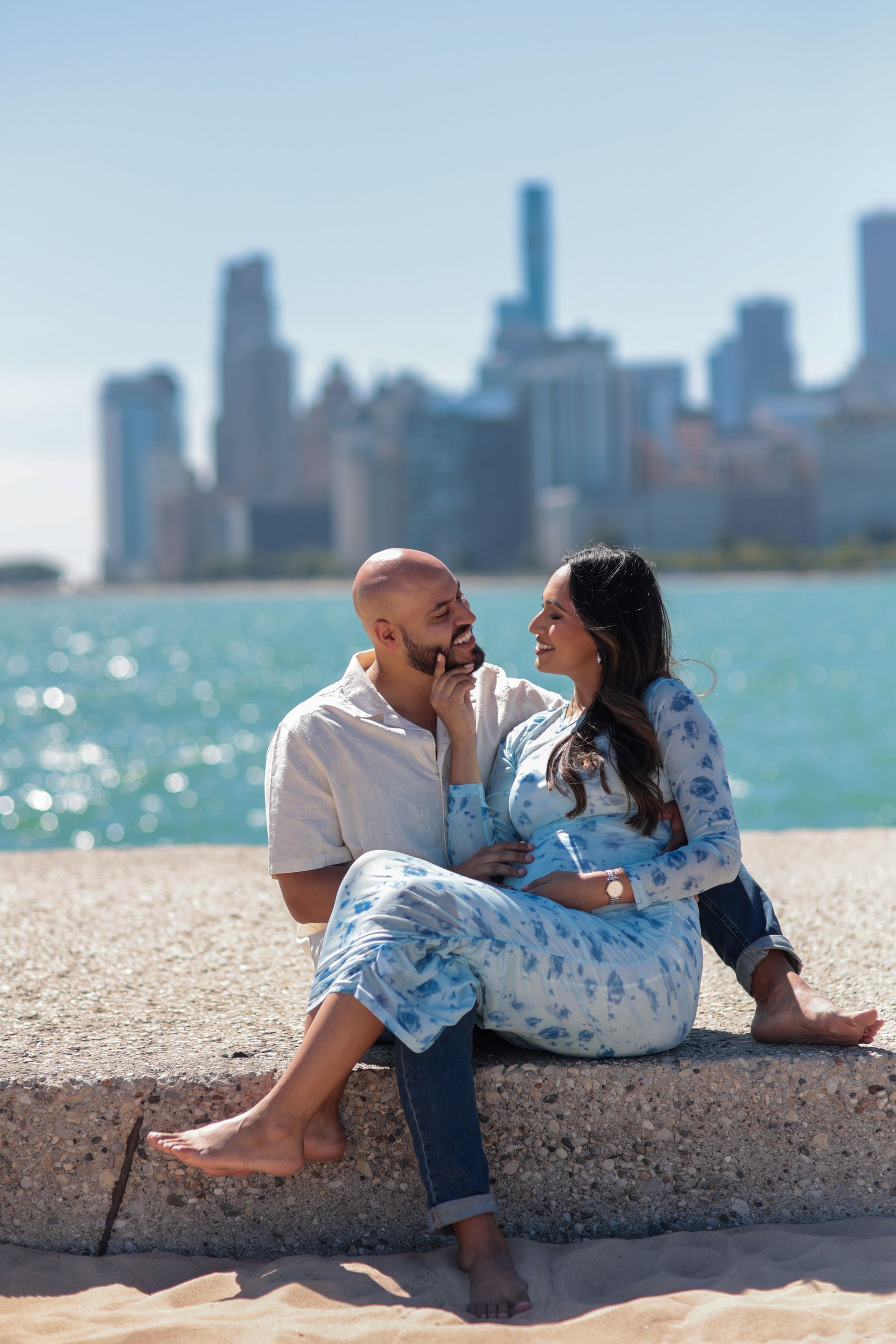 Couple sits by water, Chicago skyline backdrop. Woman pregnant, touching man's face, both smiling. Blue sky, sunny day.
