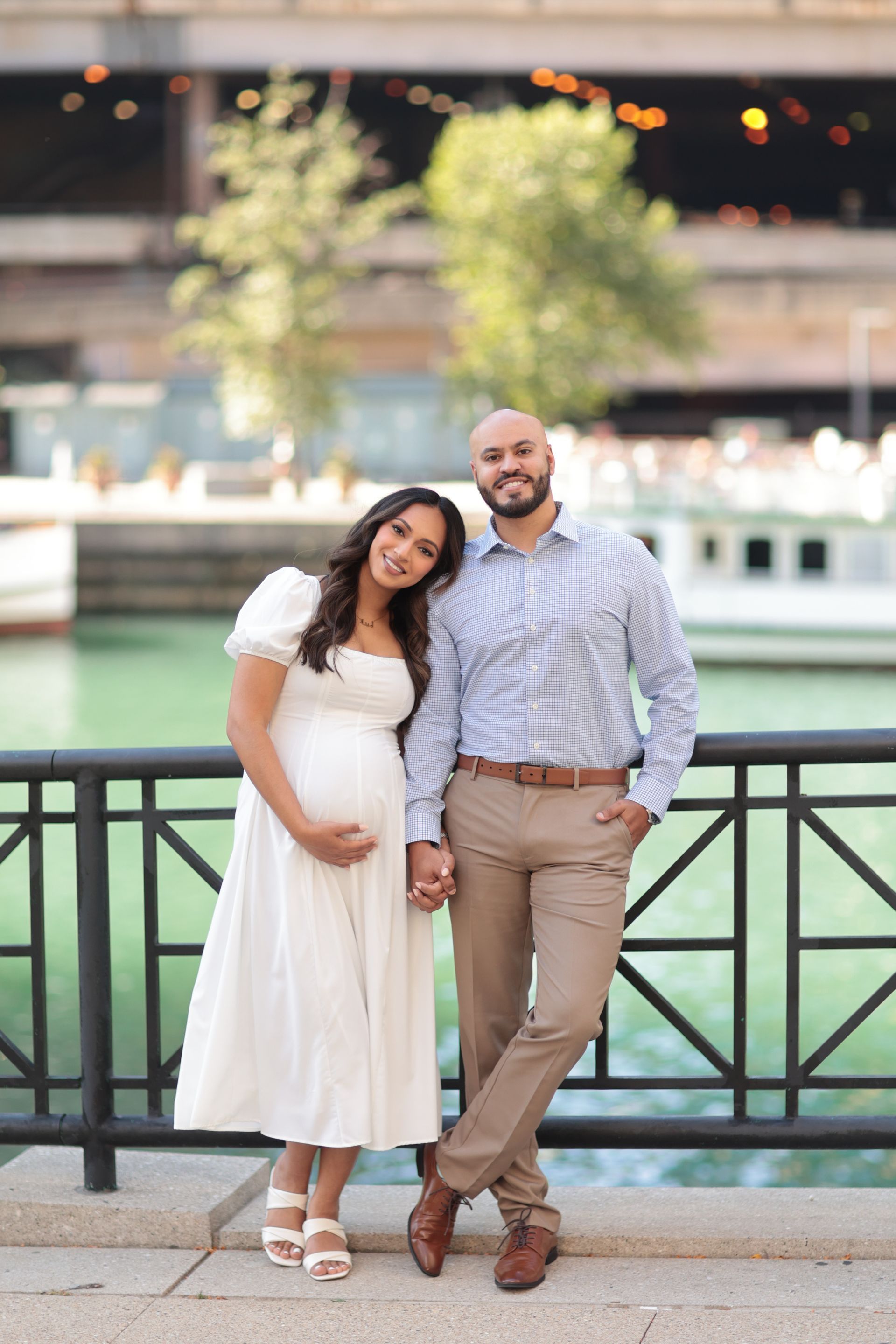 Pregnant couple poses by a river, woman in white dress leans on man in blue shirt, holding hands.