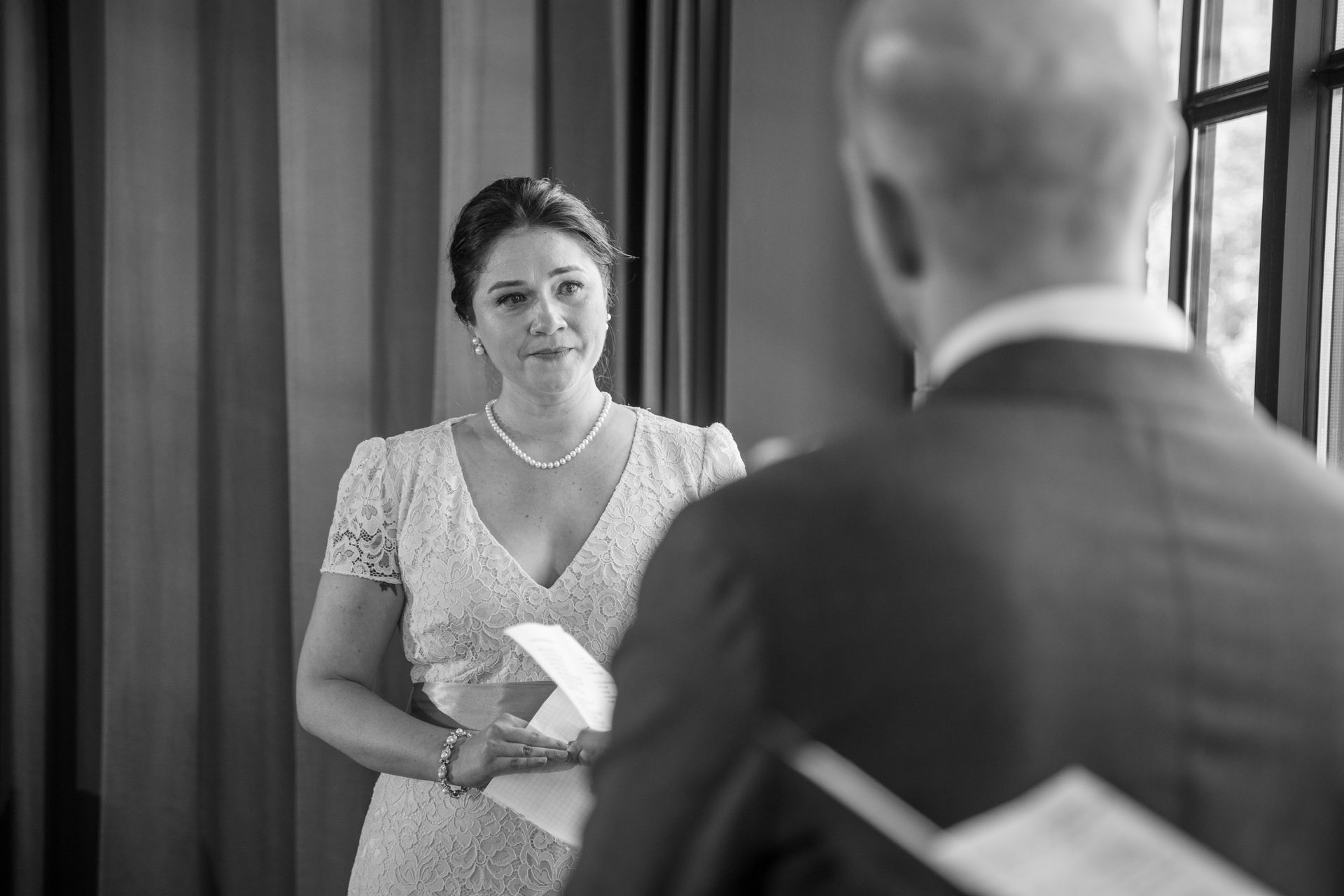 Woman in a formal dress, holding papers, looks at a person facing away from the camera. Indoor setting.
