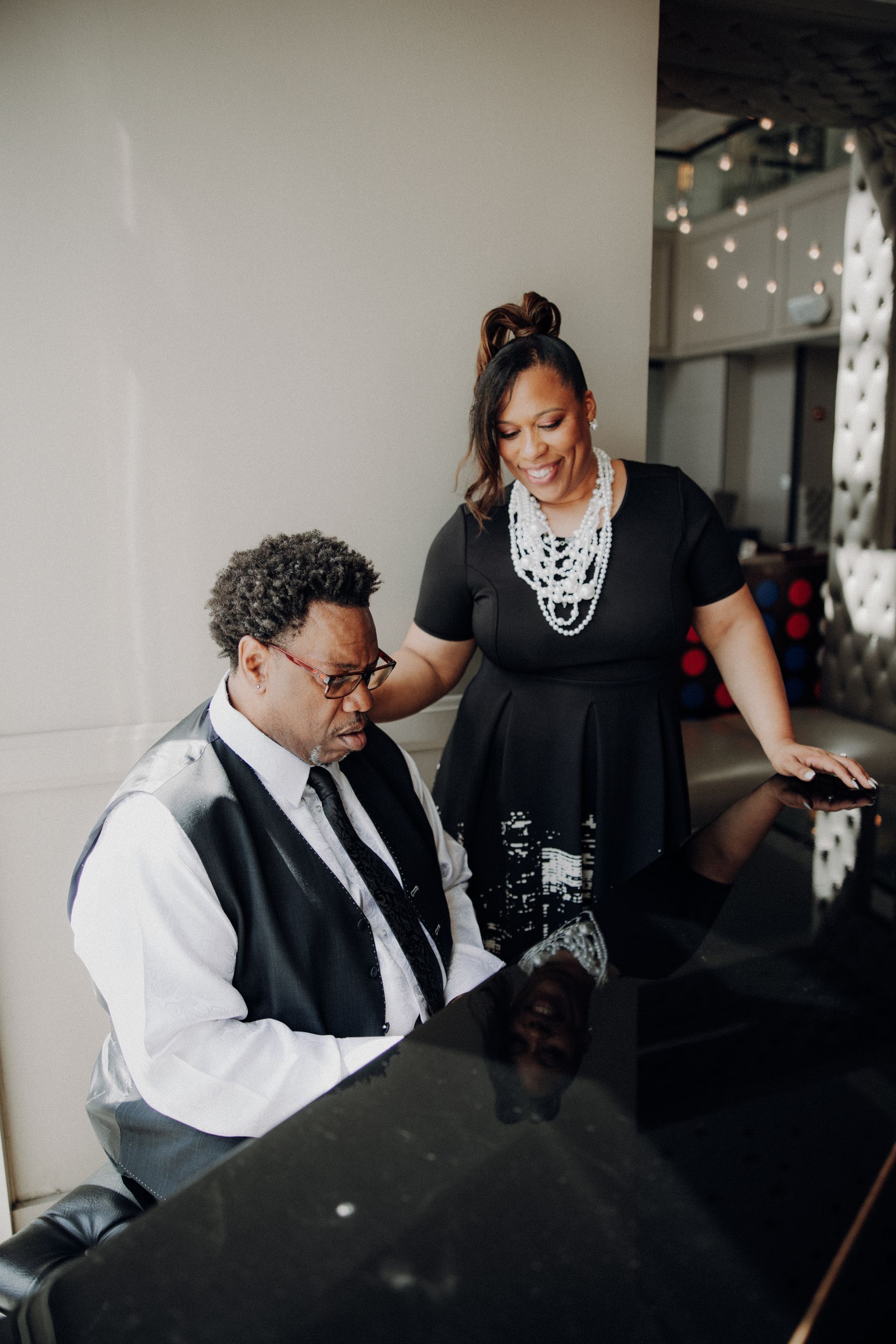 Woman smiles, touches man playing piano in a well-lit room. He wears a vest, she wears a dress and necklace.