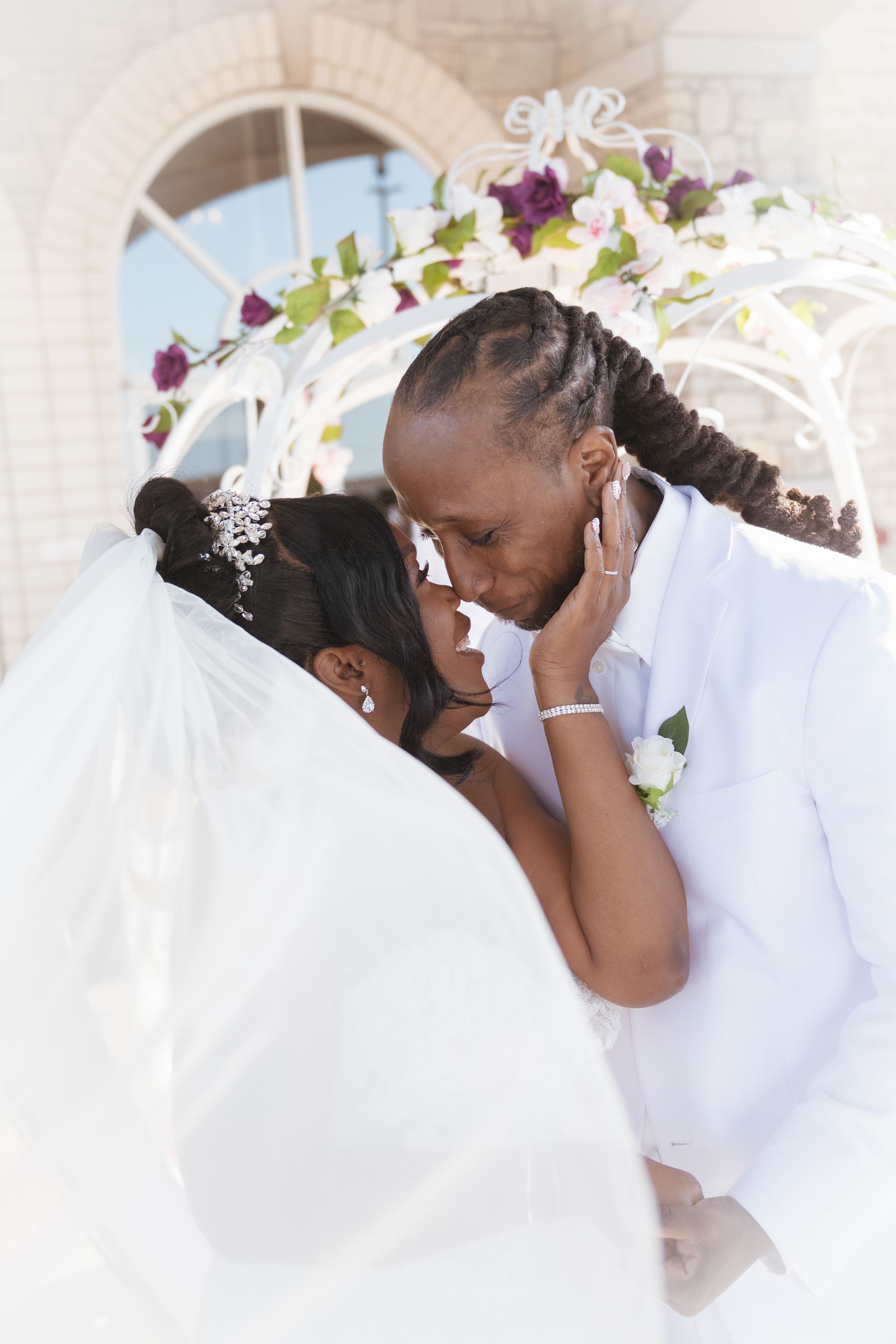 Two people embrace at wedding, veil and white attire, arch with floral decorations.