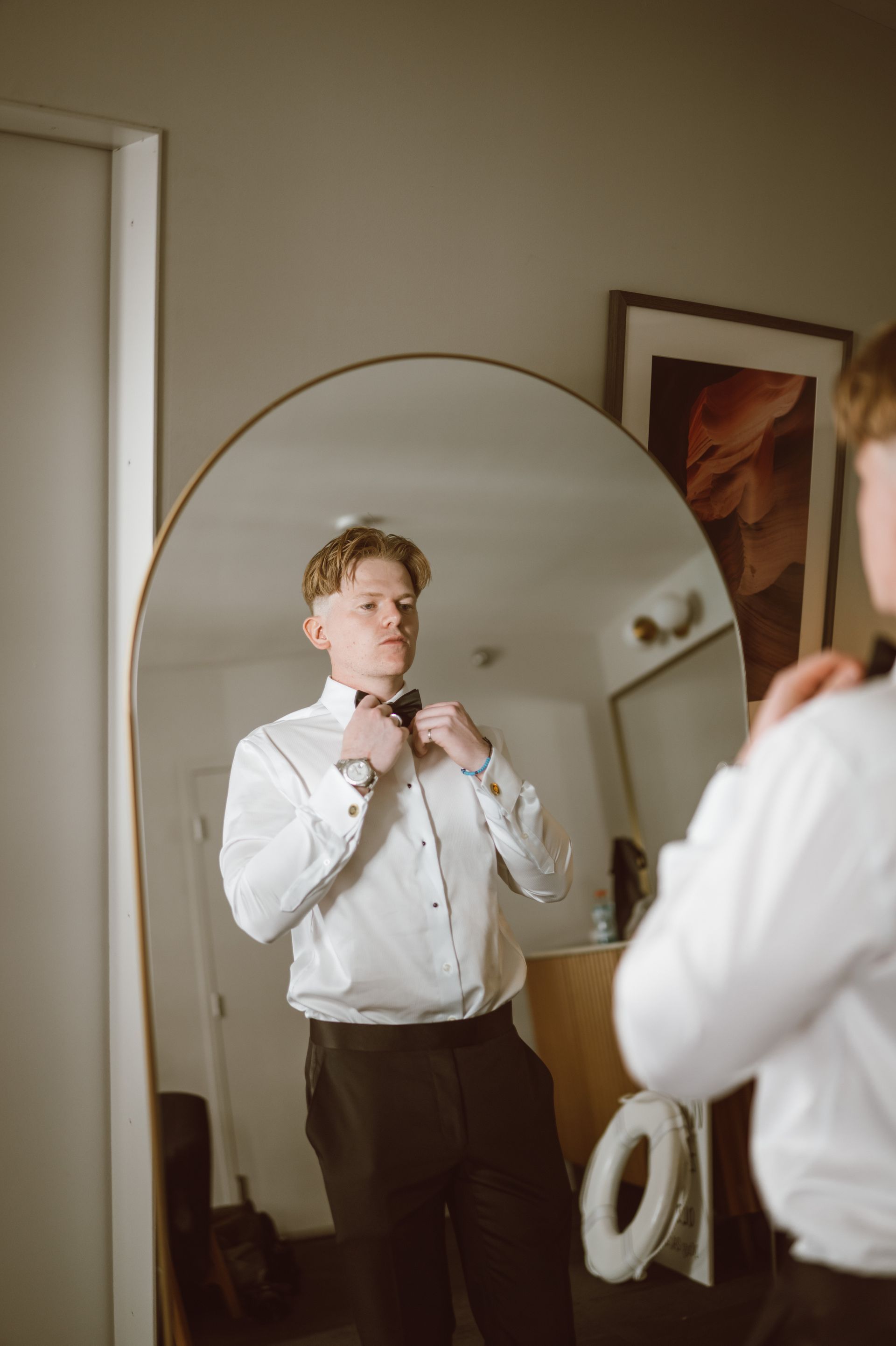 Man in white dress shirt and black pants adjusting bowtie in a mirror.