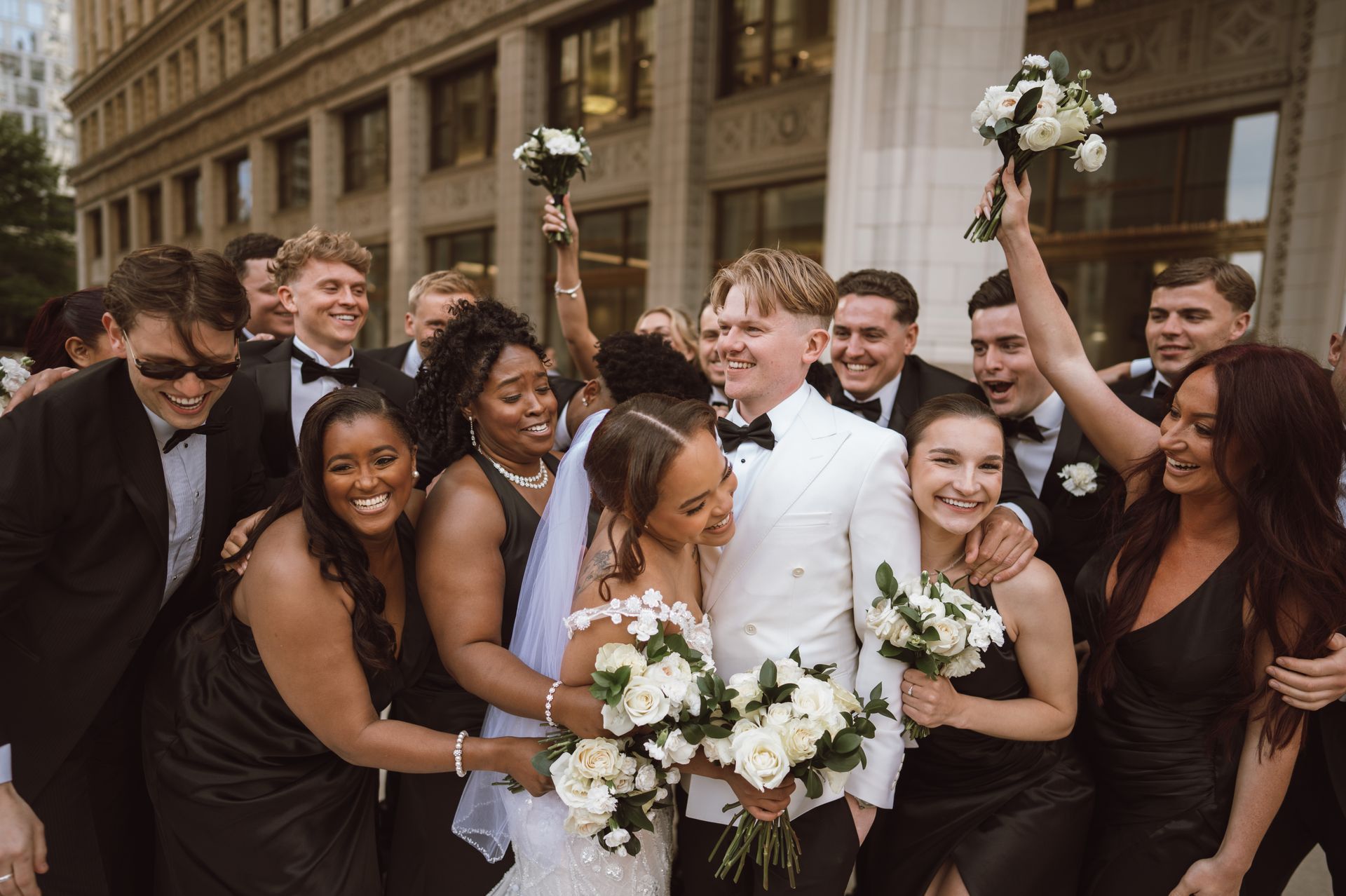 Wedding party celebrates outdoors. Couple embraces, surrounded by friends in formal attire, holding bouquets.
