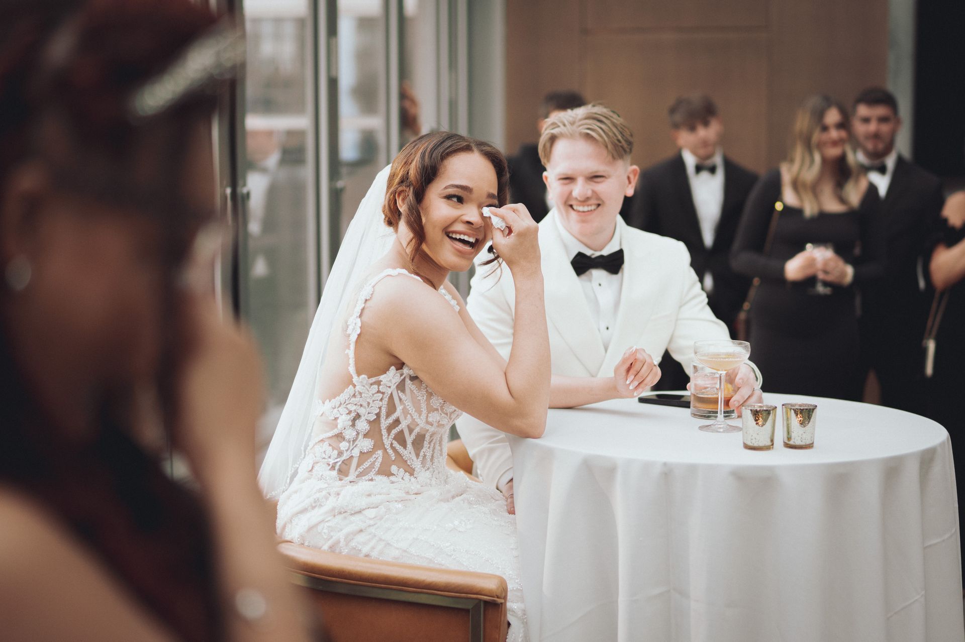 Bride and groom at a wedding reception. Bride in white dress smiles. Groom in white suit smiles. Guests in background.