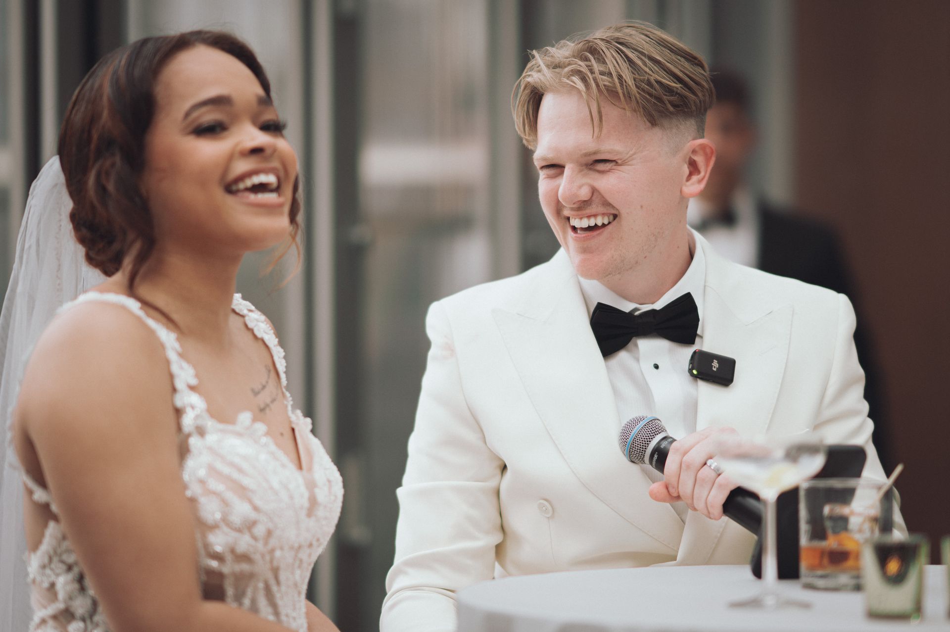 Bride and groom laughing at a reception. The groom holds a microphone; a martini and other drinks are on the table.