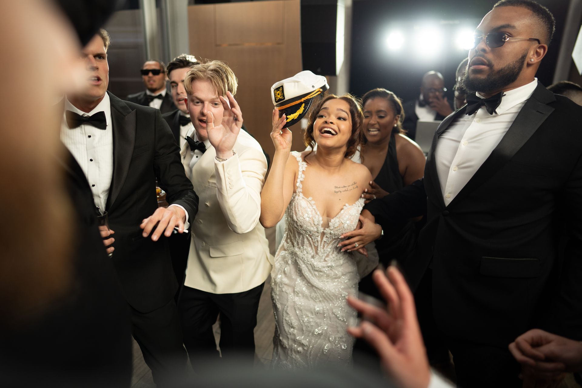 A joyful woman in a sparkly gown celebrates with friends, holding a captain's hat. Others wear tuxedos.