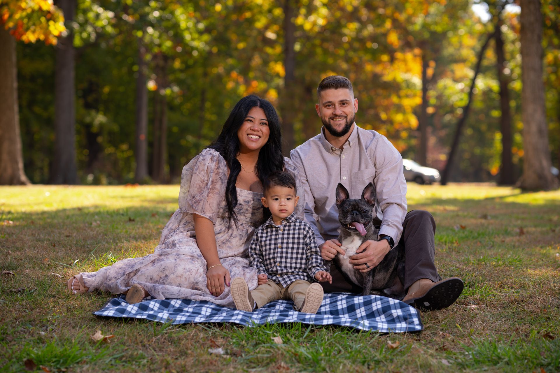 Family of three and dog sit on a blanket in a park; woman, child, man, and French bulldog look at the camera.