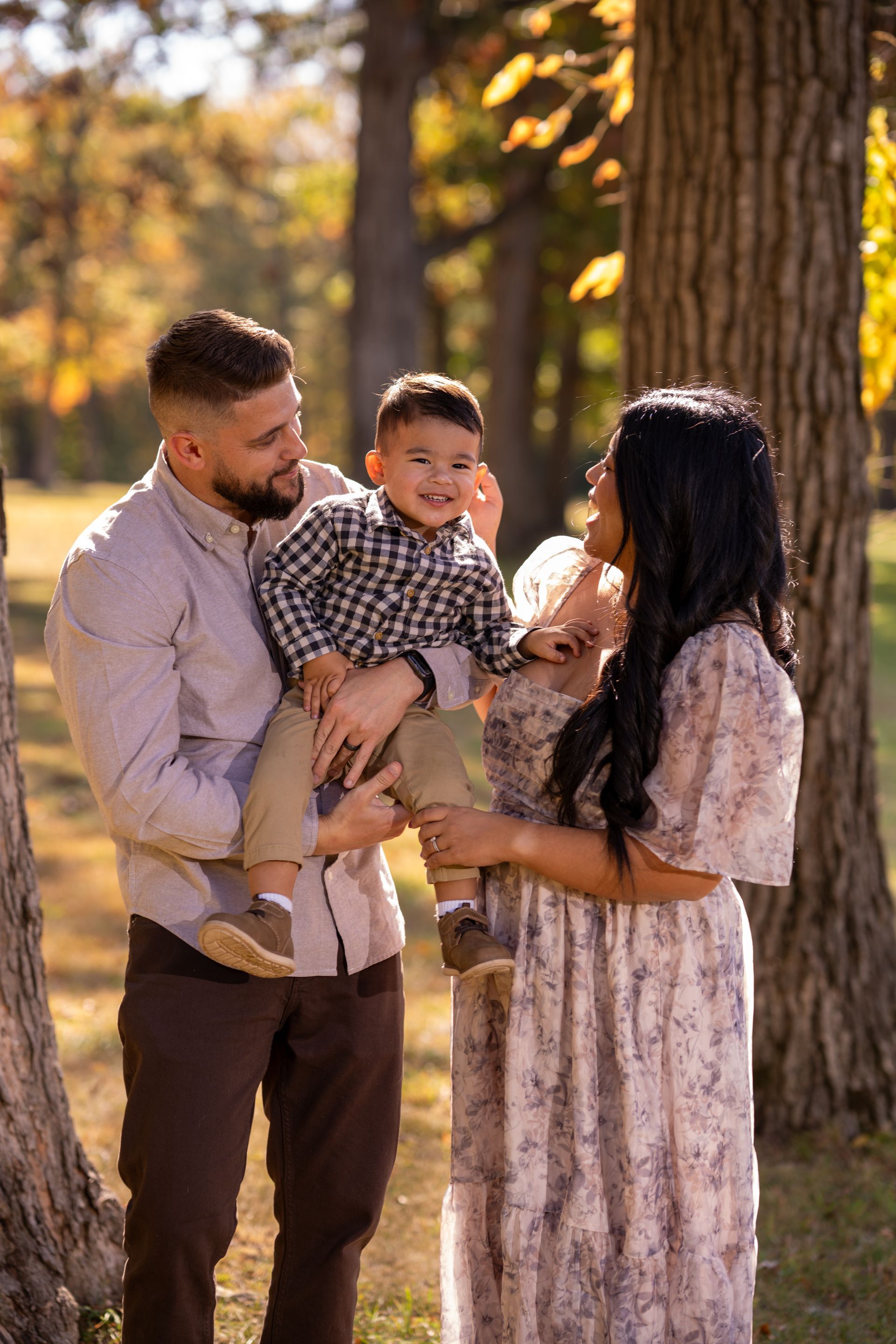 Family of three smiling in a park, man holding toddler. Woman beside them in a floral dress. Autumnal setting.