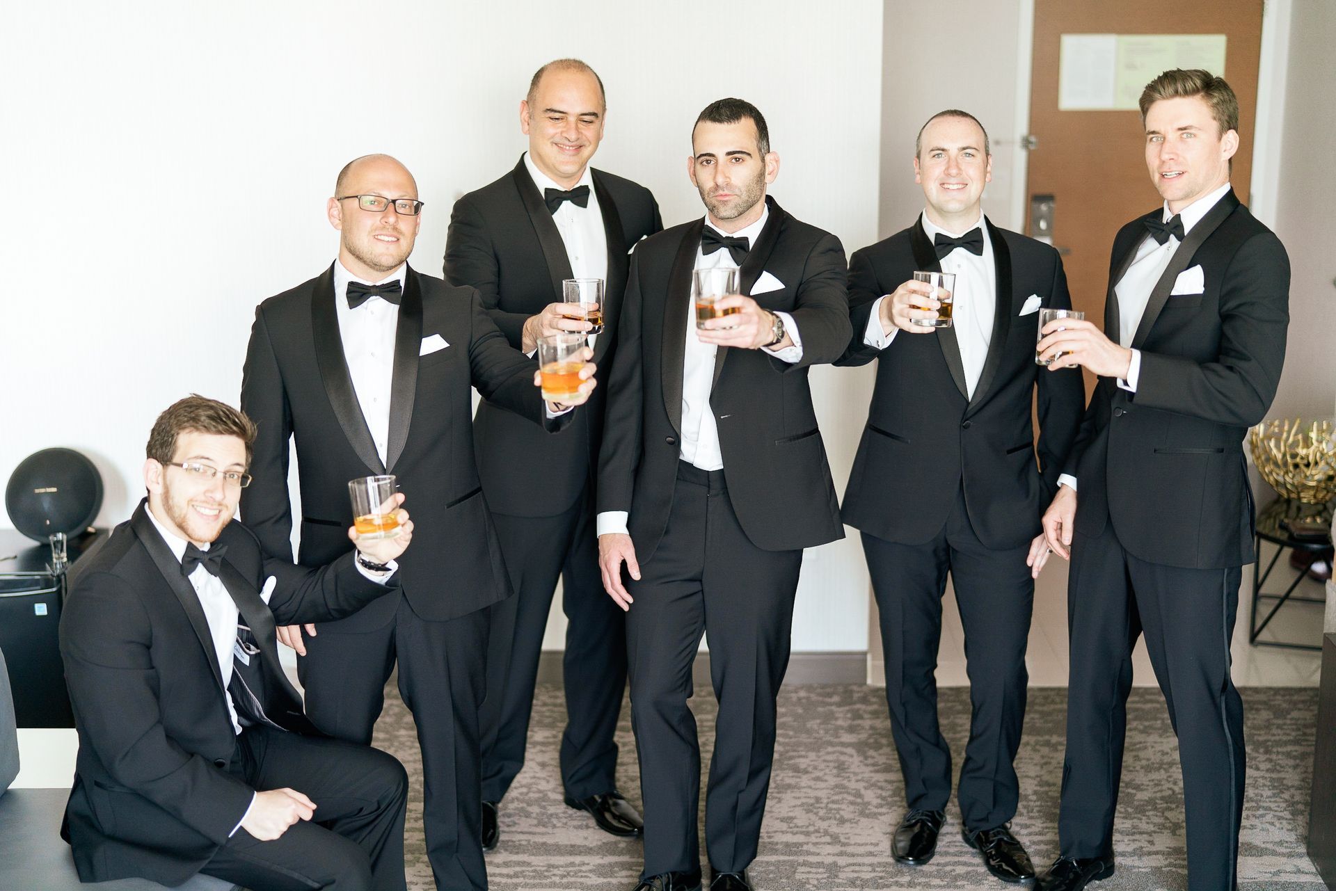 Men in tuxedos hold drinks, posing together in a bright room.