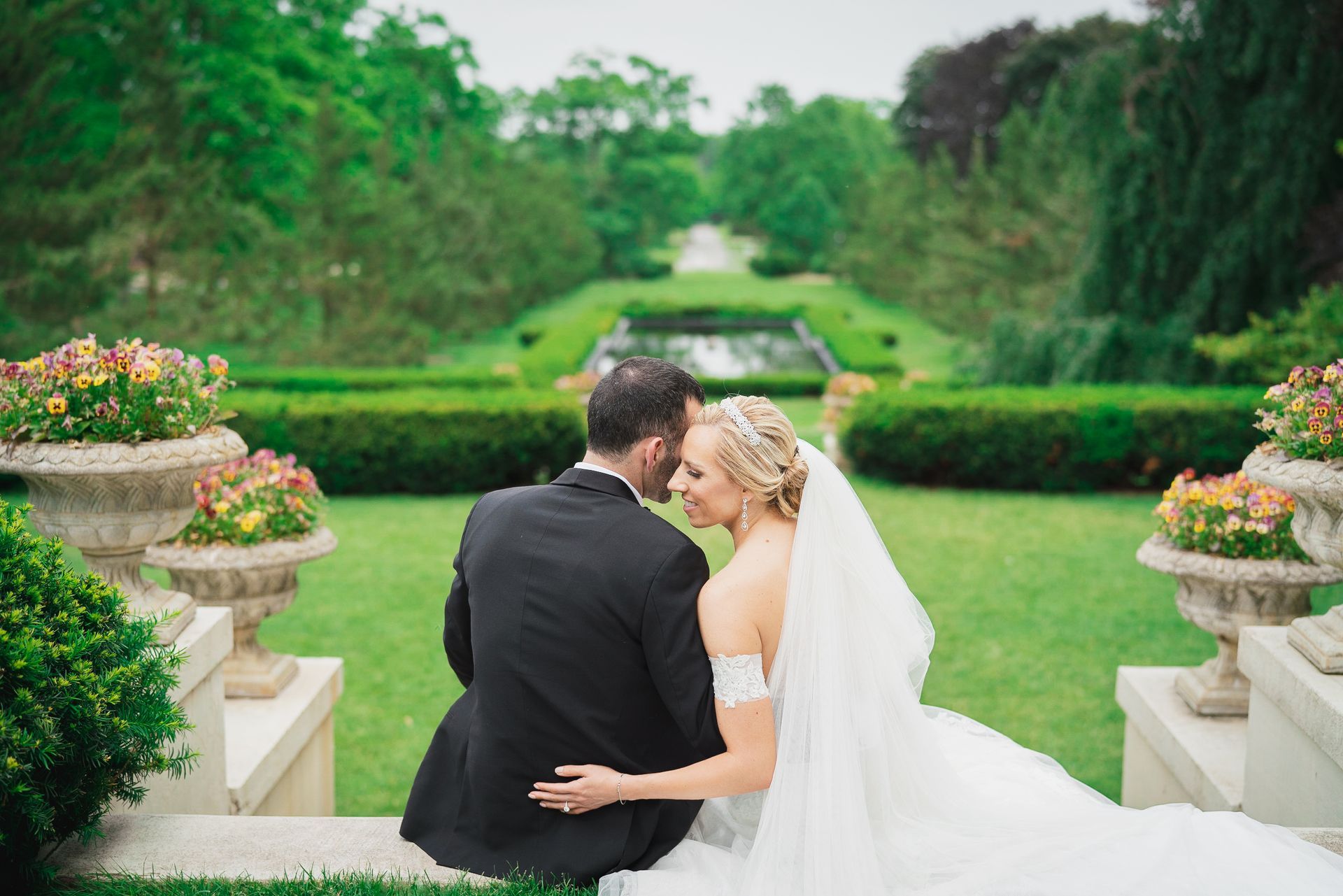 Bride and groom seated on steps, embracing, with a formal garden and pond in the background.