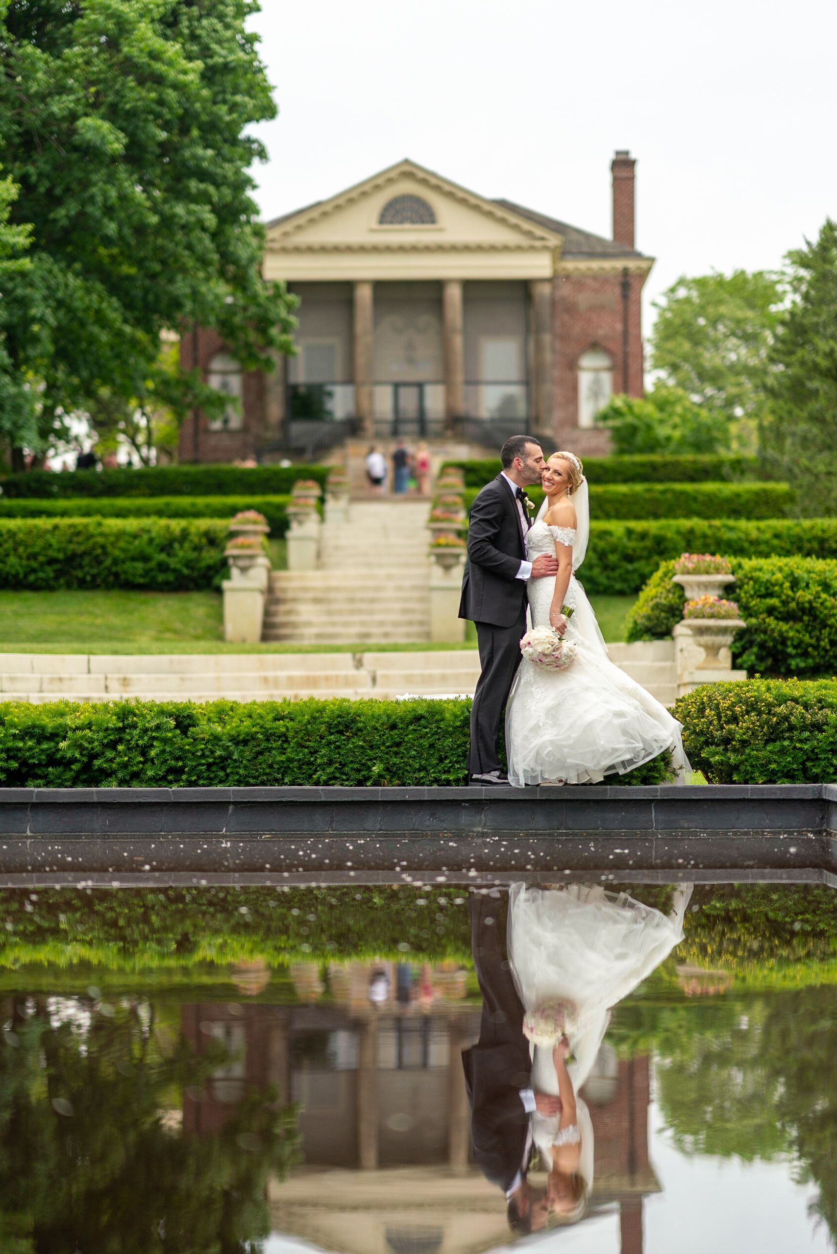 Bride and groom embrace in front of a mansion, reflected in a pond. They are smiling, he is kissing her.