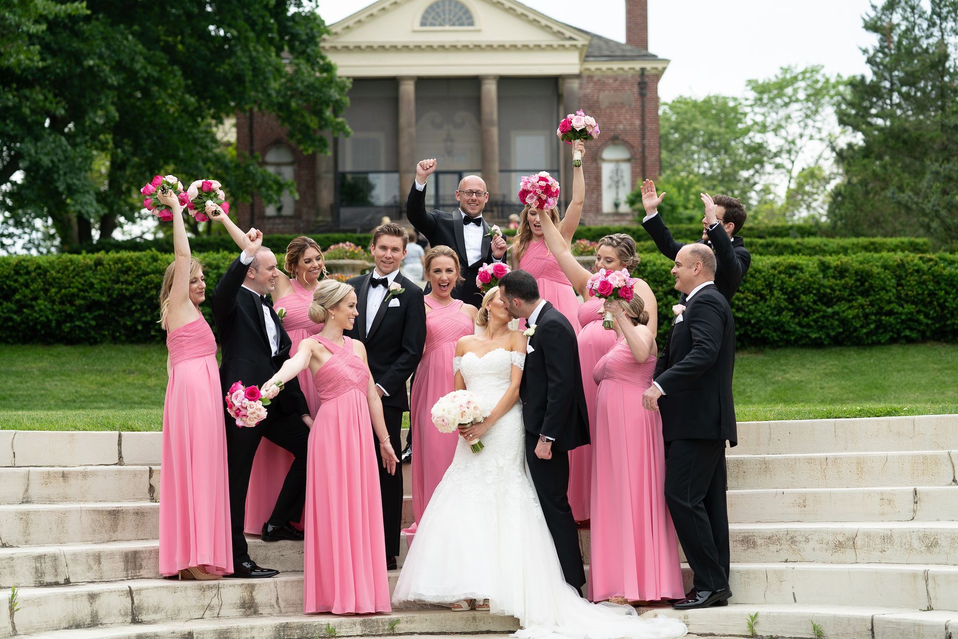 Wedding party celebrating on outdoor steps. Bride and groom kissing; bridesmaids in pink dresses.