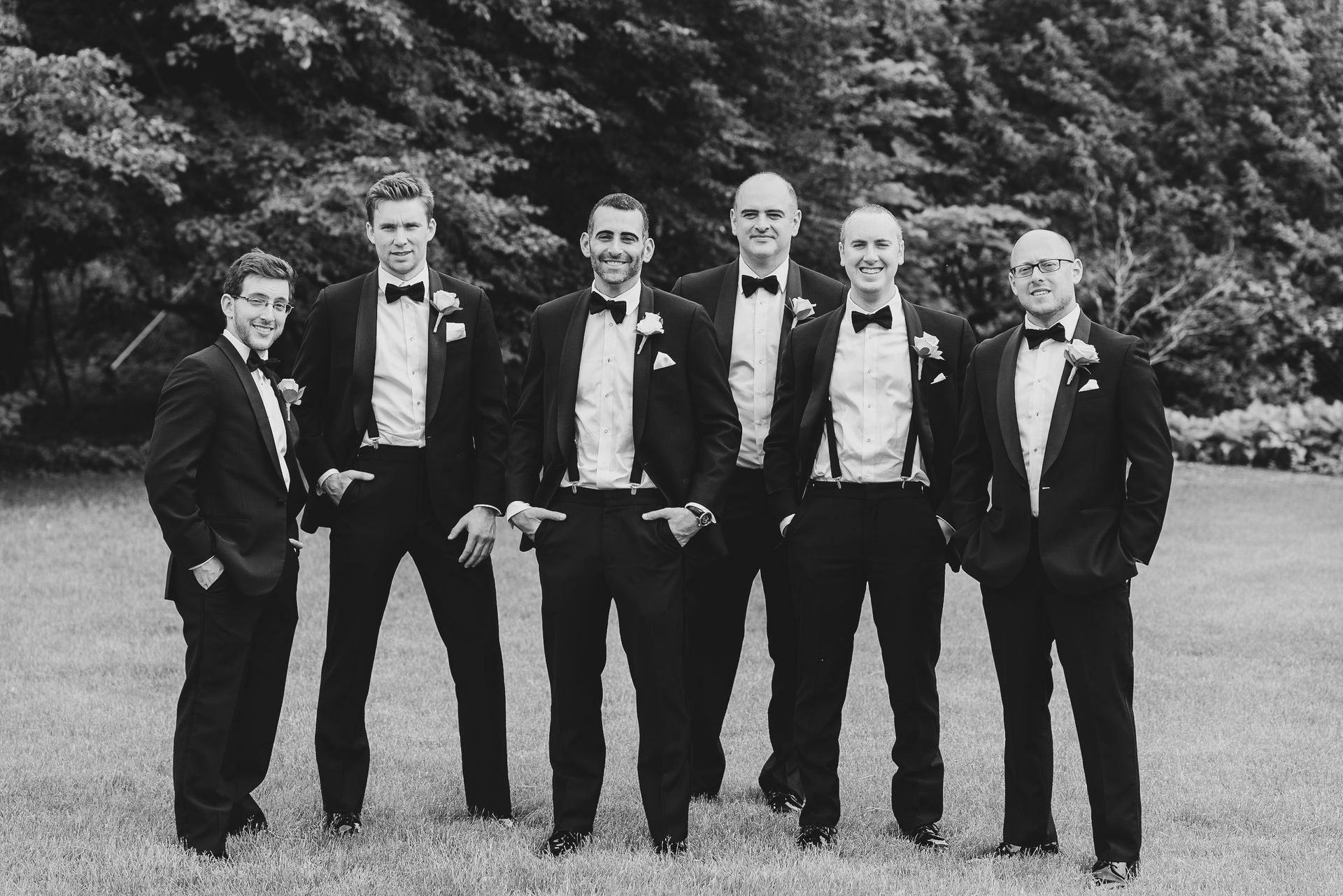 Men in tuxedos stand on grass, some with suspenders, smiling, in front of trees.