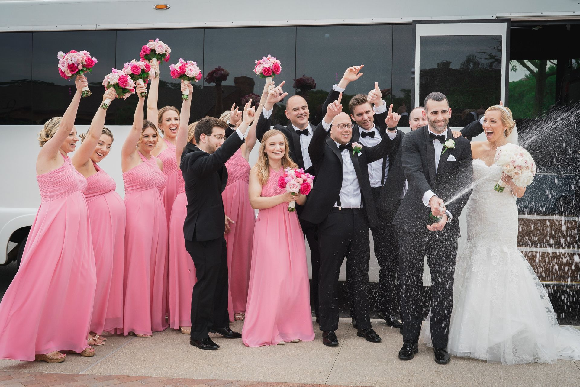 Wedding party celebrates, popping champagne near a white bus. Bridesmaids wear pink, groomsmen in black tuxedos.
