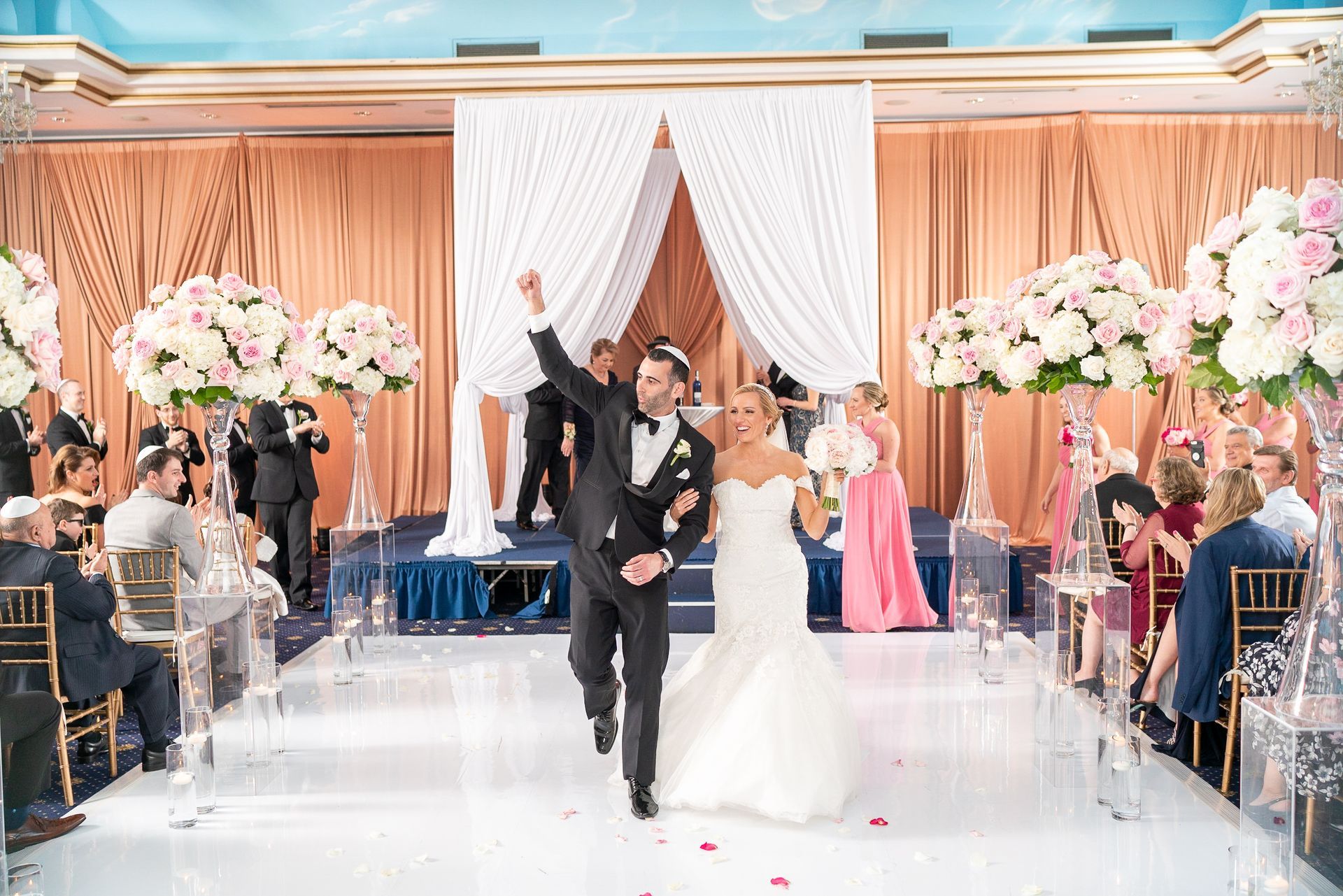 Newlyweds celebrate after a wedding ceremony. The groom raises his arm. Bride smiles, holding bouquet. Ceremony in a ballroom.