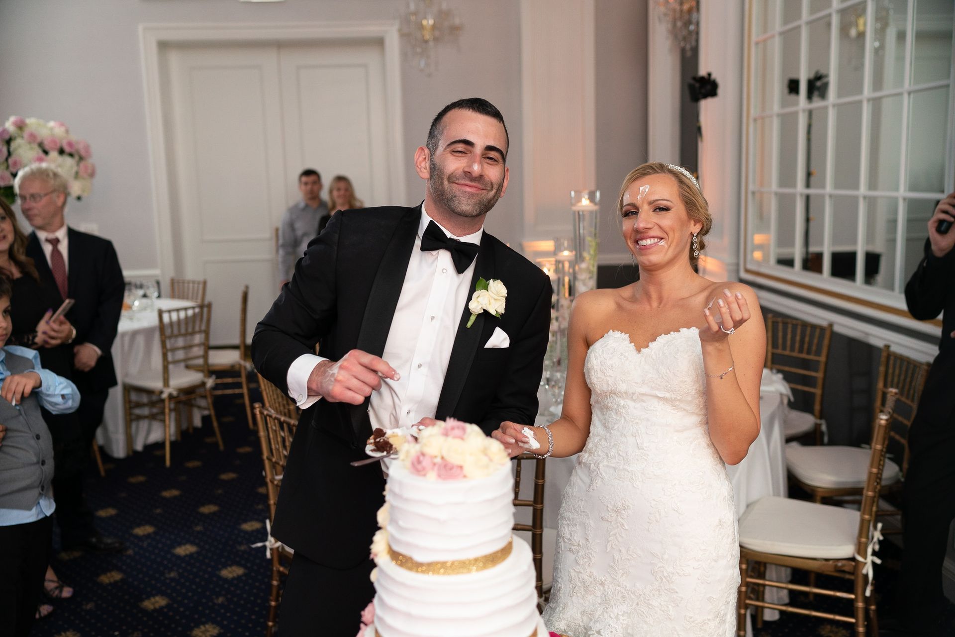 Bride and groom cutting a tiered wedding cake; smiles and happy expressions, indoor reception.