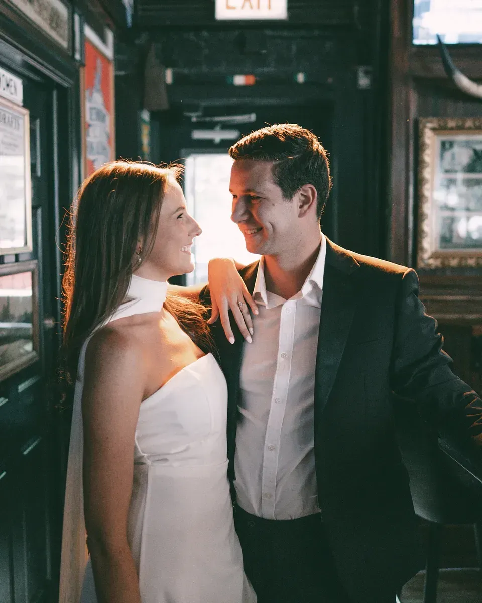 Couple smiling at each other. Woman in white dress, arm on man's shoulder. Man in dark suit, white shirt. Inside a pub.