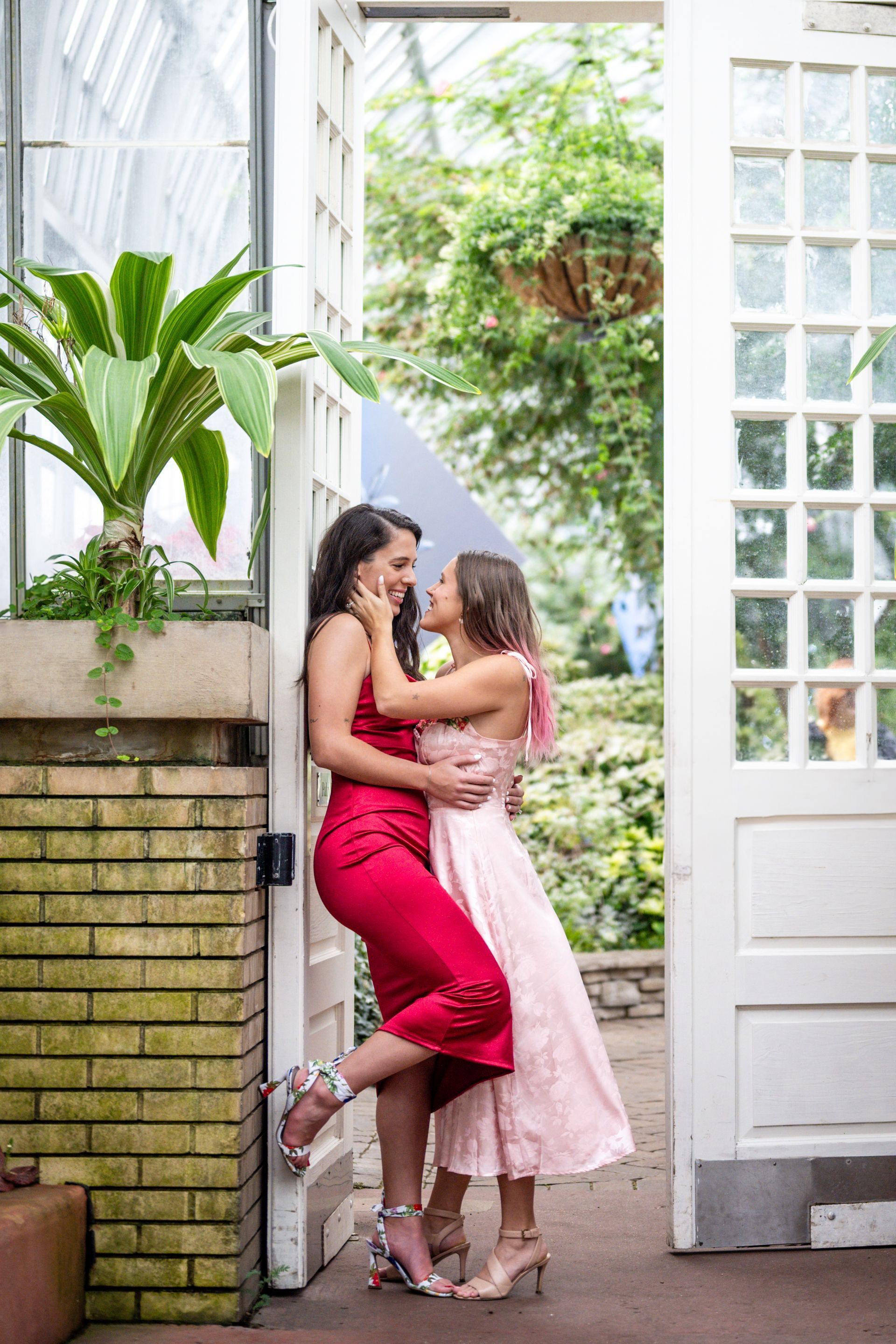 Two women embrace in a doorway. One wears red, the other pink. Lush greenery surrounds them.
