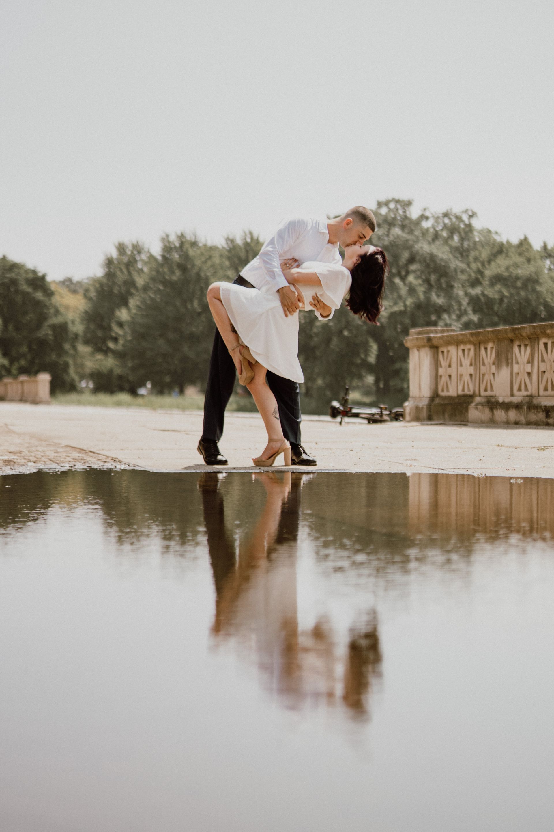 Couple kissing, reflected in a puddle. The man in white shirt and dark pants leans back, woman in white dress arches. Outdoors.