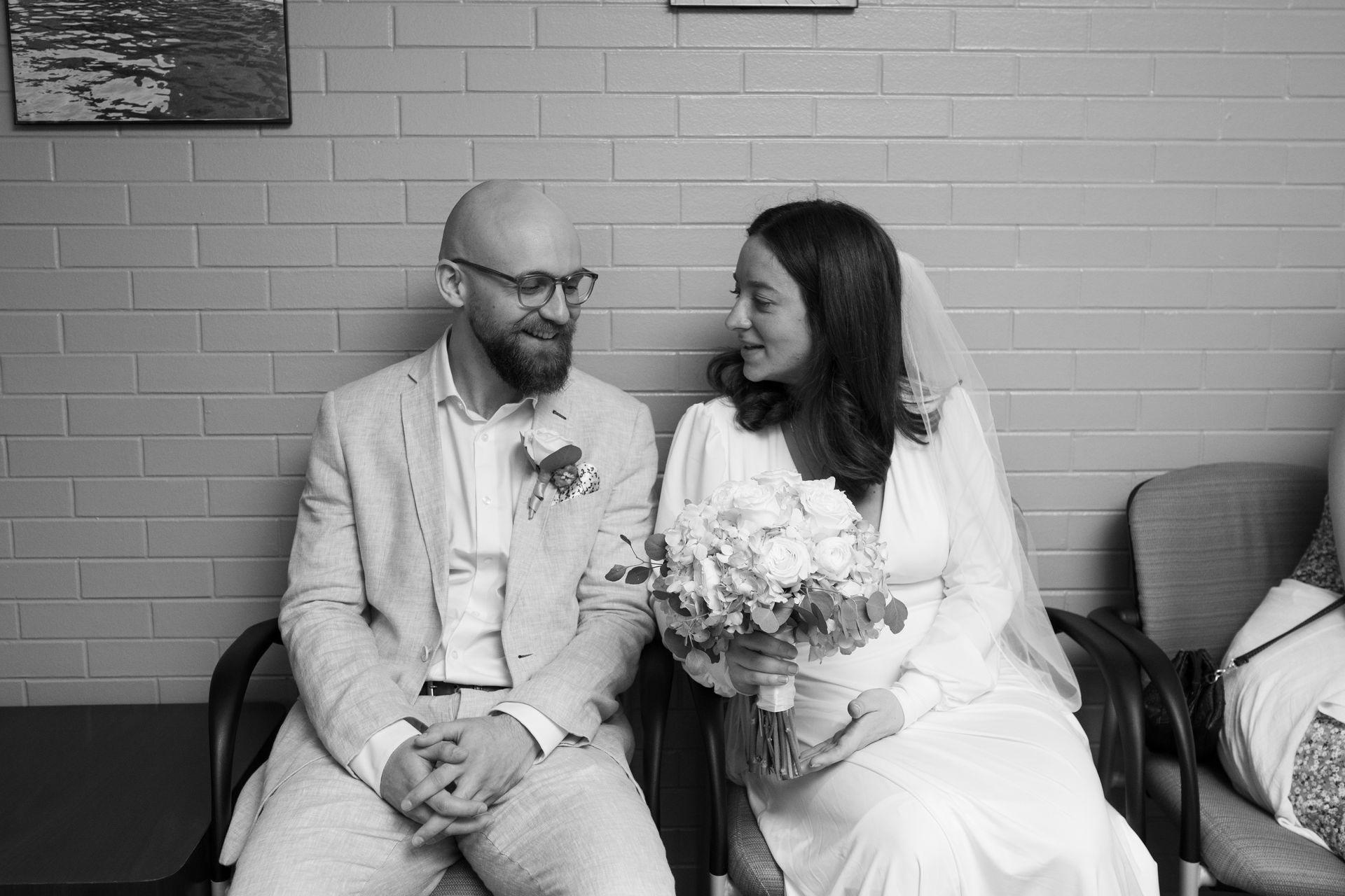 Bride and groom sitting close, smiling at each other. He wears a suit, she holds a bouquet, both in a waiting room.