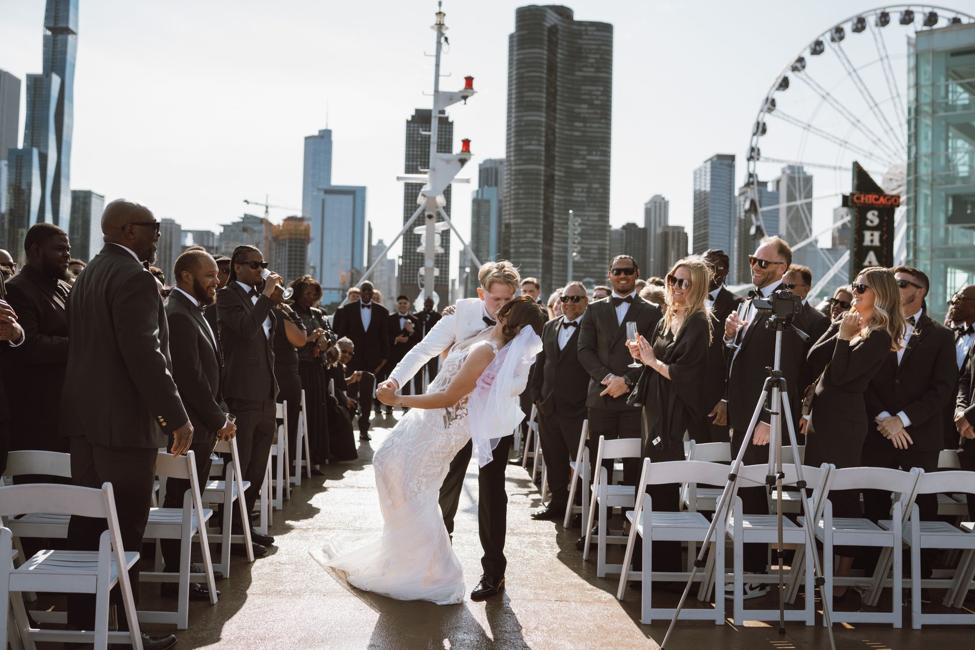 Newlyweds dip to kiss, surrounded by wedding party, city skyline and Ferris wheel visible.