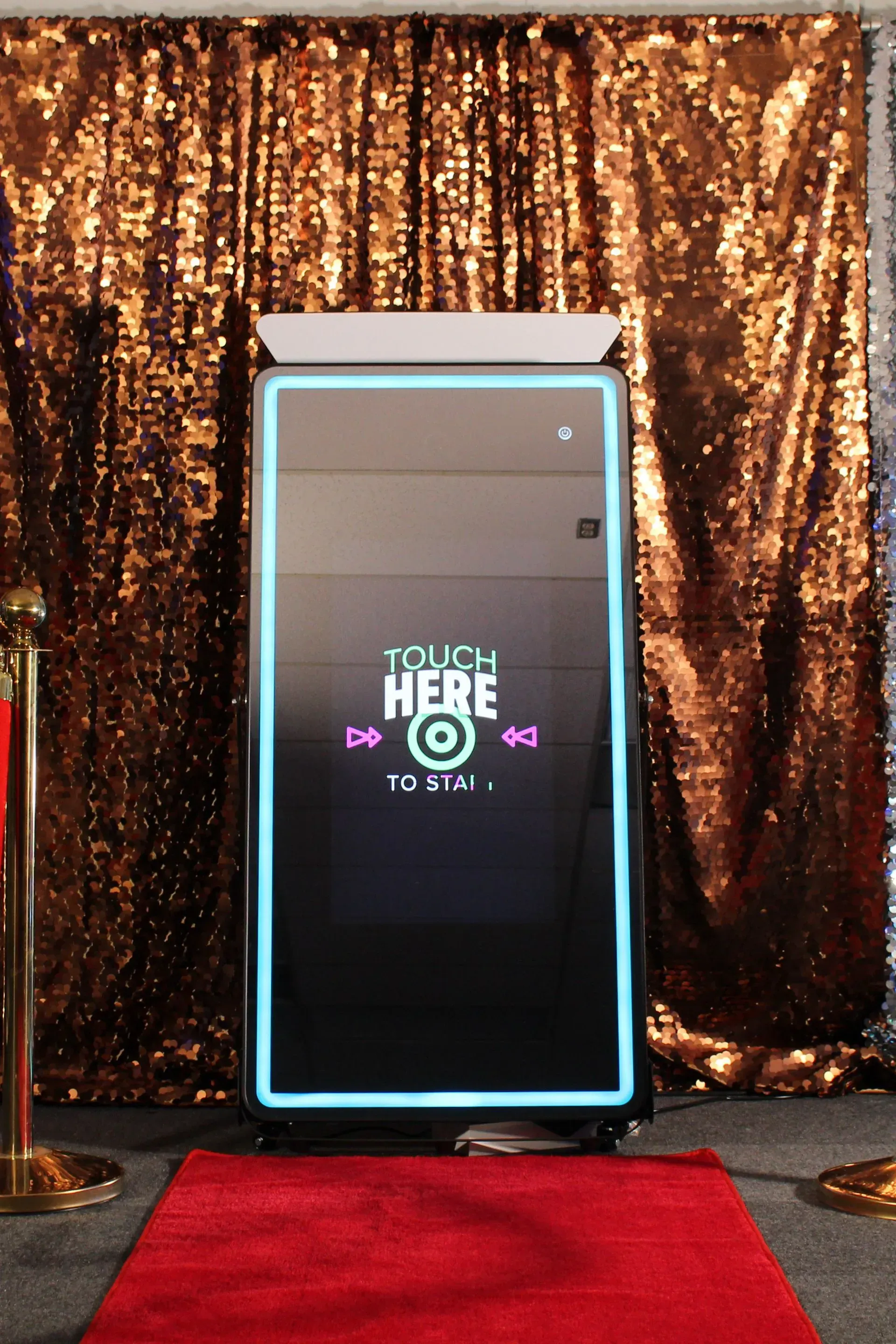 Photo booth with touch screen, neon border, on red carpet, gold sequin backdrop, ready to start.