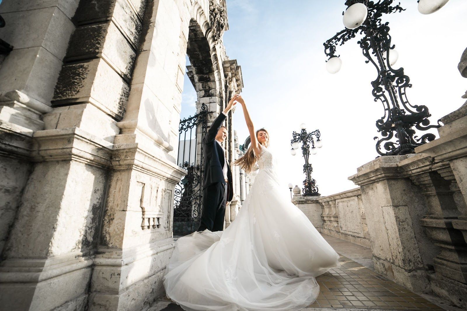 Bride and groom dancing, celebrating wedding day on stone balcony, under ornate street lamps.