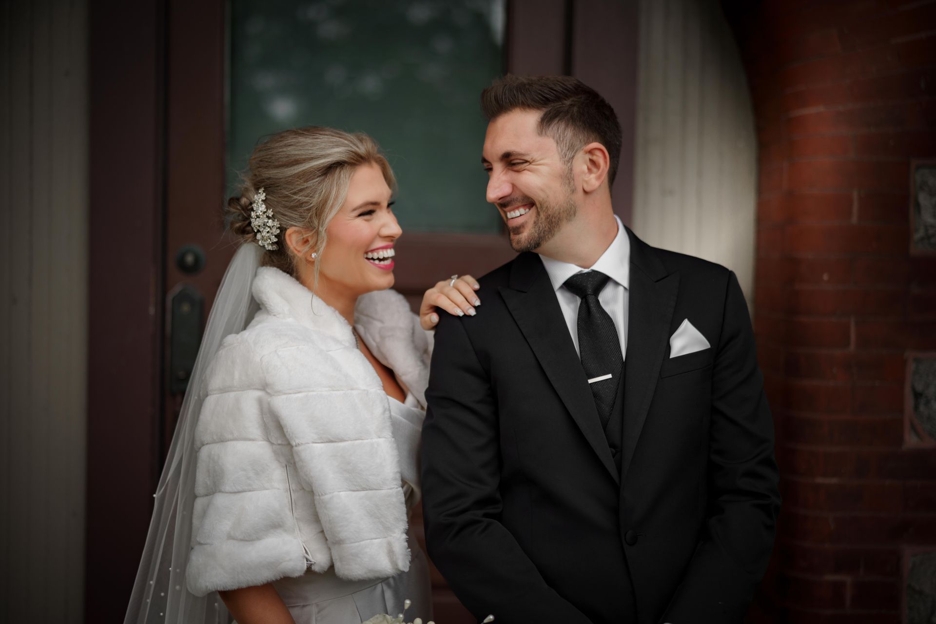 Bride and groom laugh, bride wearing fur wrap, groom in black suit, in front of a building.