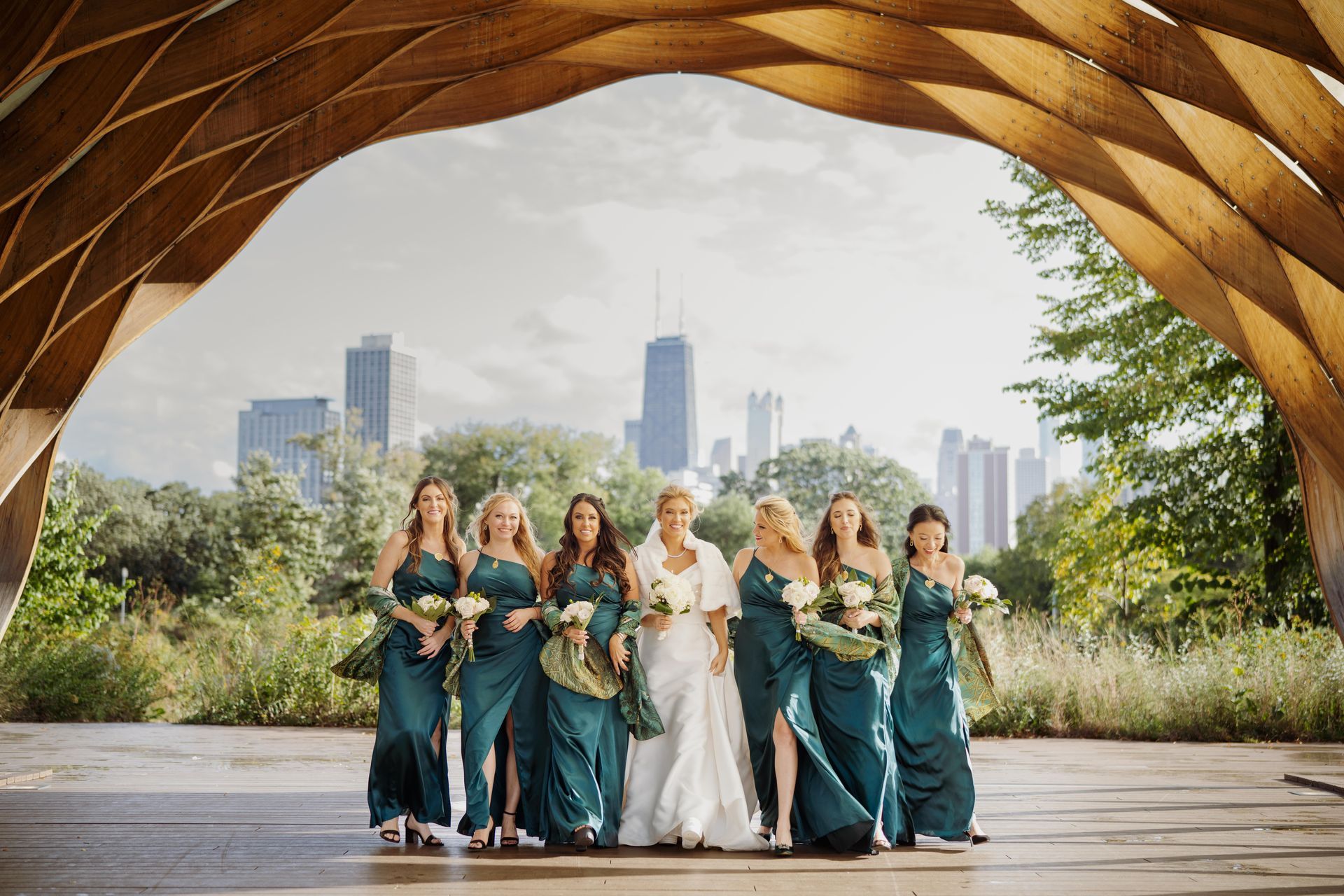 Bride and bridesmaids in teal dresses pose beneath a wooden archway with a city skyline backdrop.