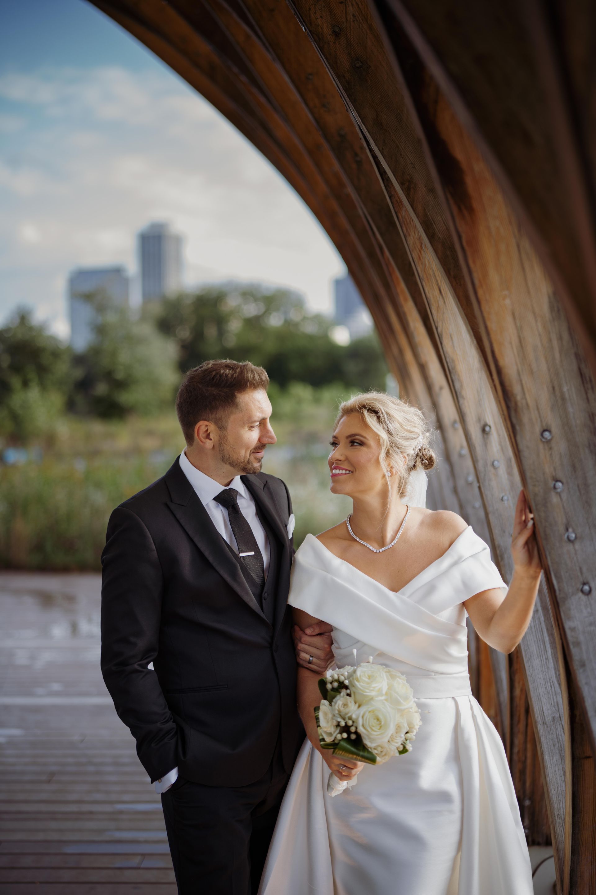 Couple in wedding attire gazes at each other, standing under a wooden archway with a city skyline in the background.