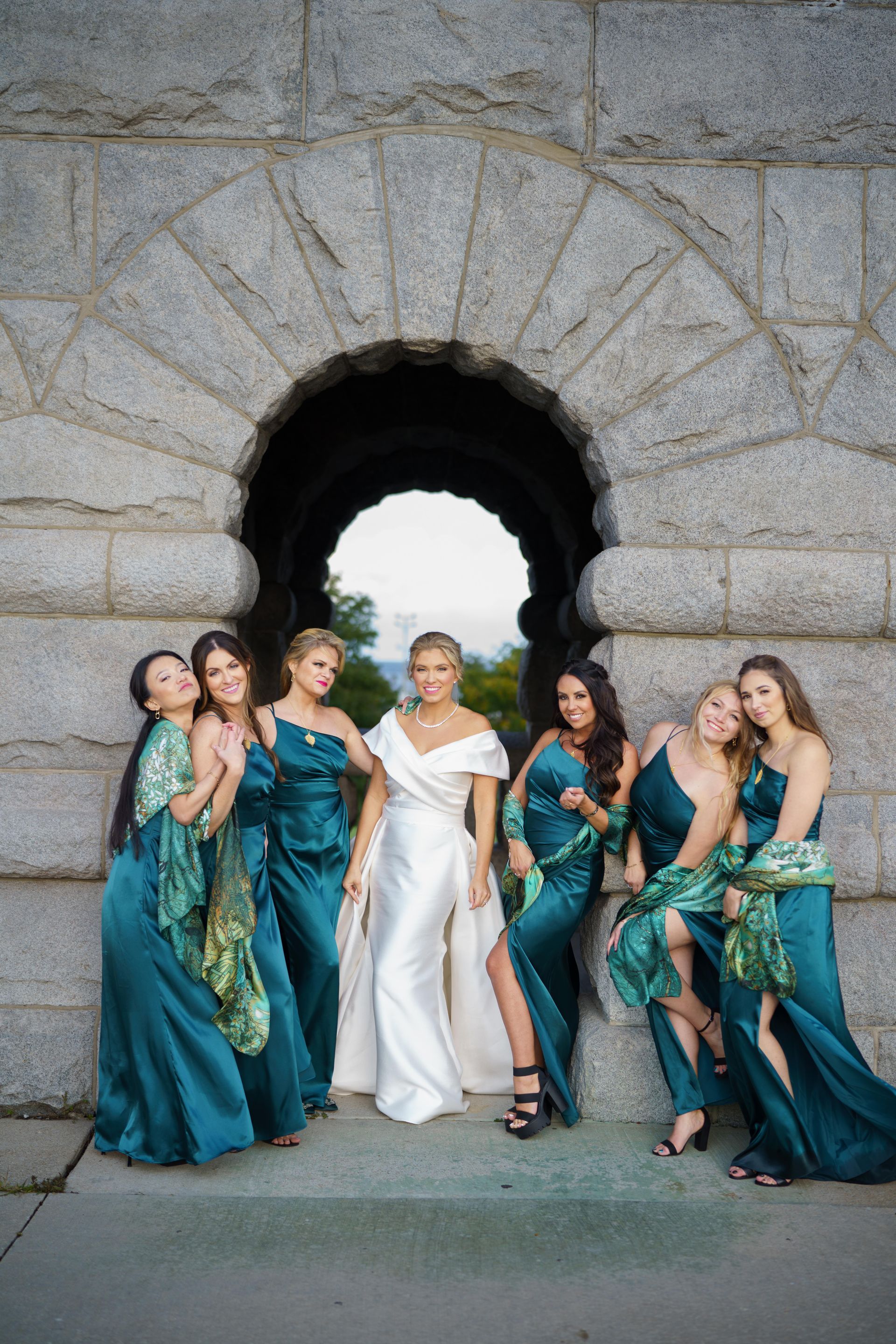 Bride in white gown poses with bridesmaids in teal dresses under an arch.