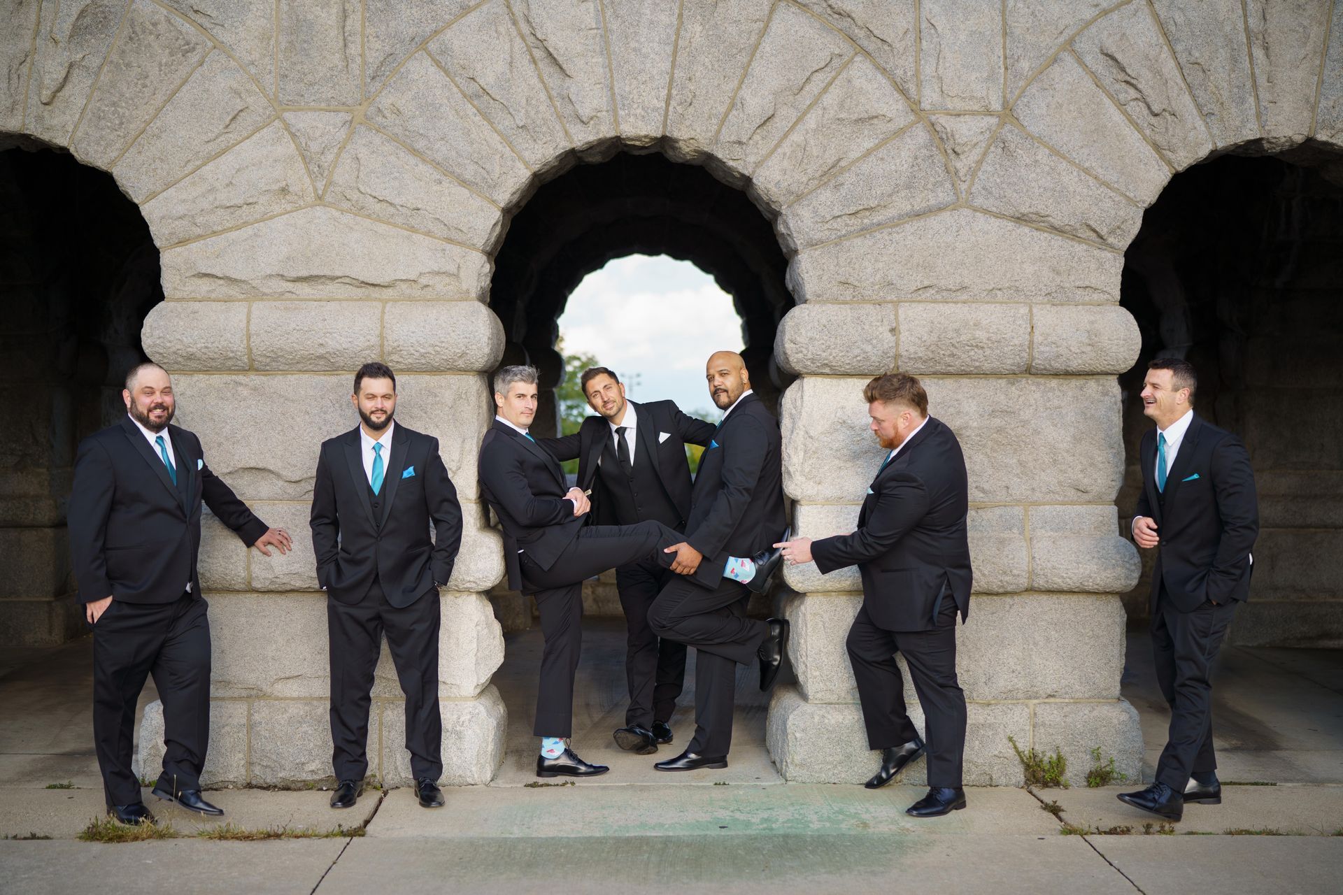 Men in suits pose playfully under an arched stone structure, some with turquoise accents.
