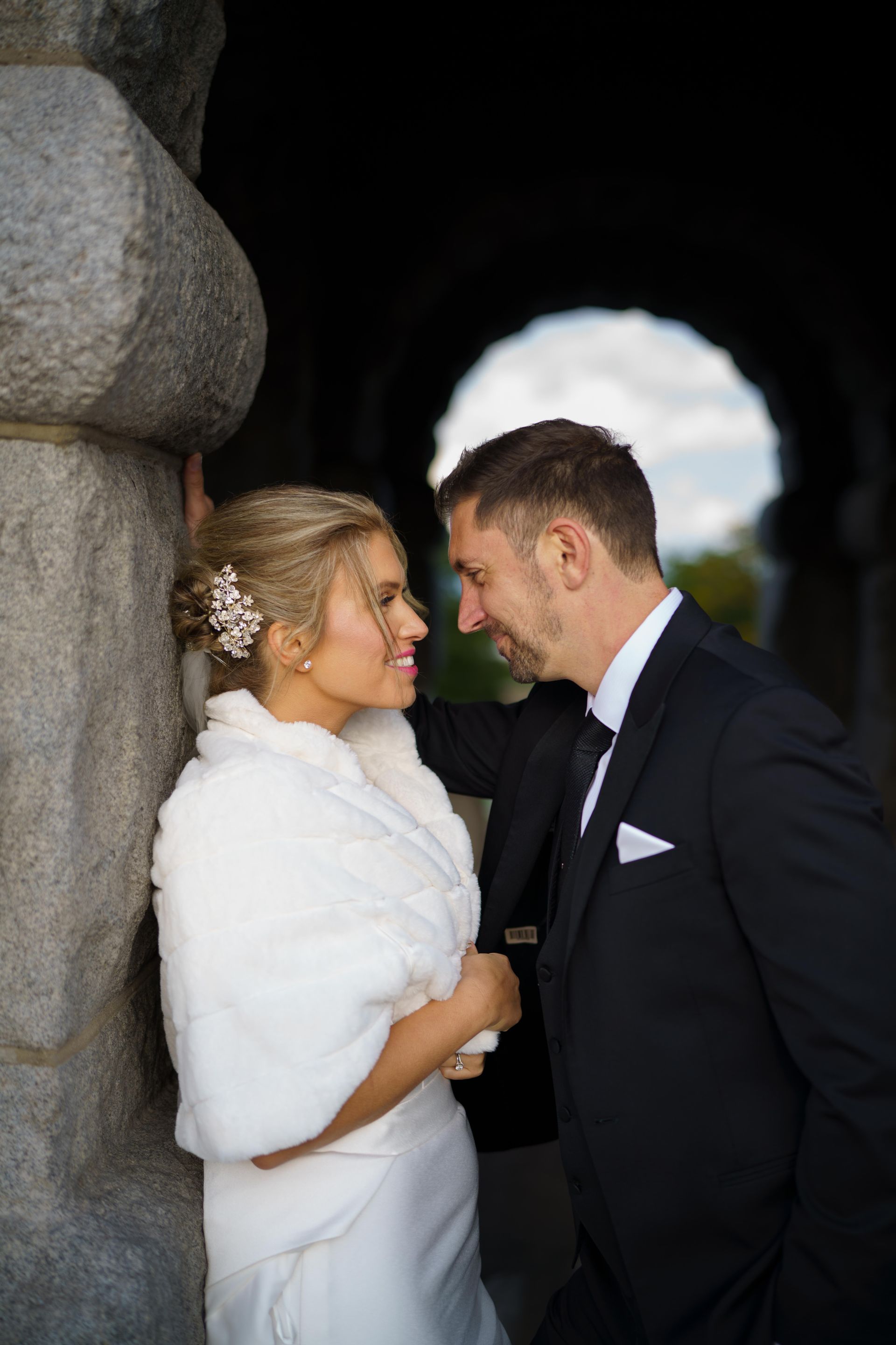 Bride and groom in formal wear gaze at each other. She leans on stone pillar, he stands beside her.