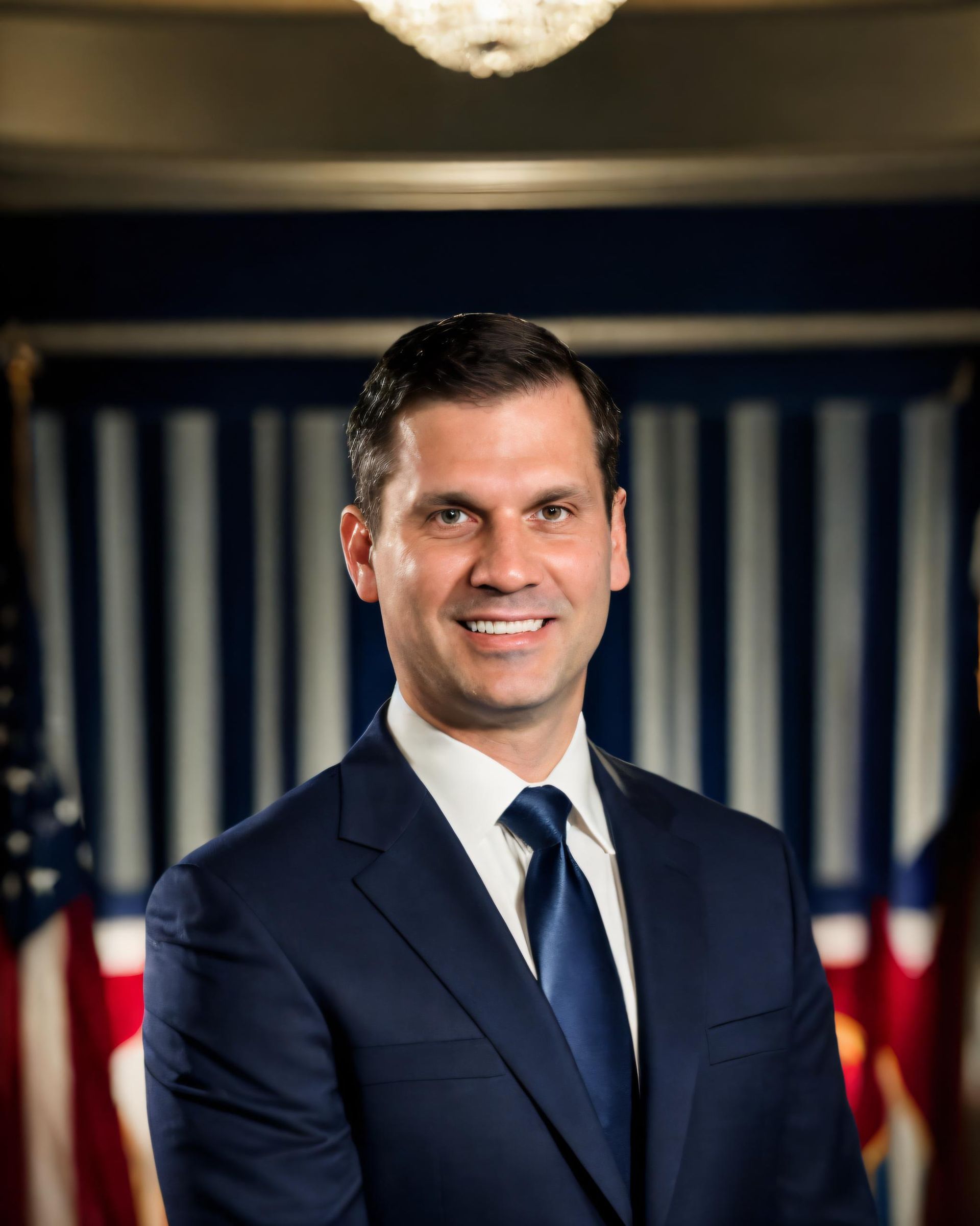 Smiling man in a navy suit and tie standing before a blurred U.S. flag backdrop