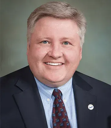 Smiling man in a dark suit, light blue shirt, and patterned tie against a gray studio background