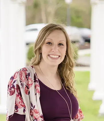 Smiling woman in a purple top and floral cardigan standing outdoors on a porch