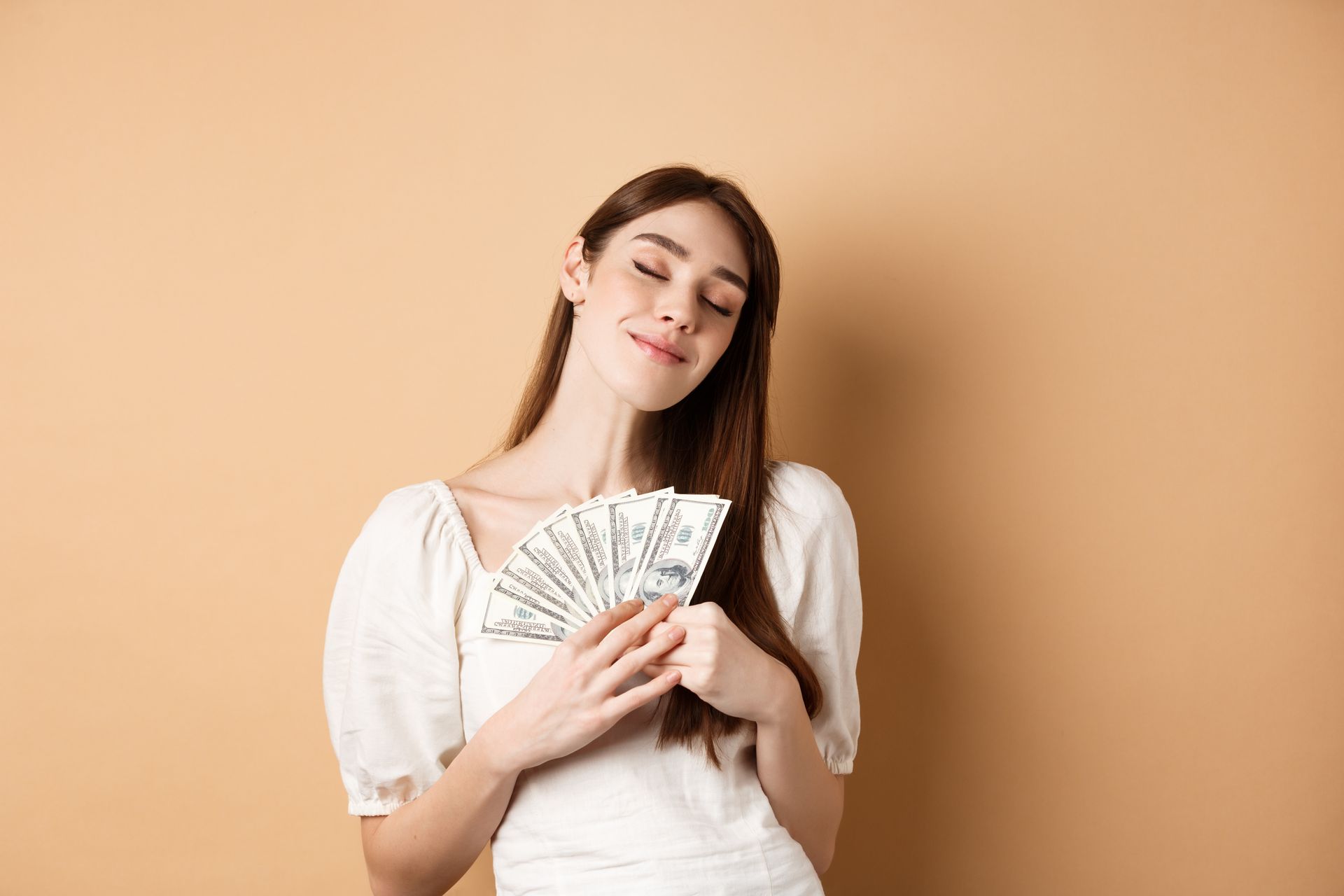Woman in white dress holding fan of money, smiling with eyes closed. Beige background.