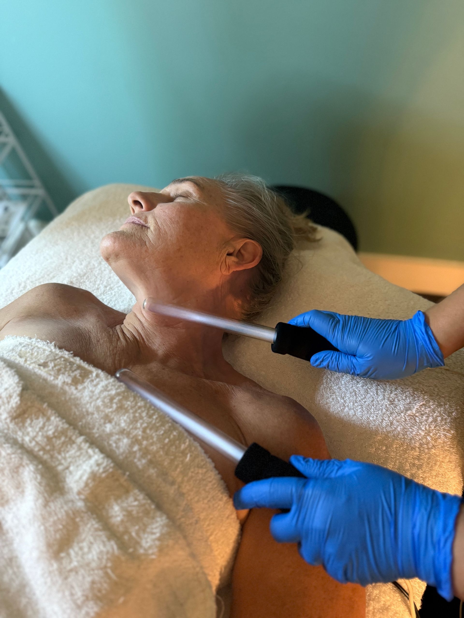 Gloved hands apply a facial treatment to a person lying on a spa bed with a towel over their chest.
