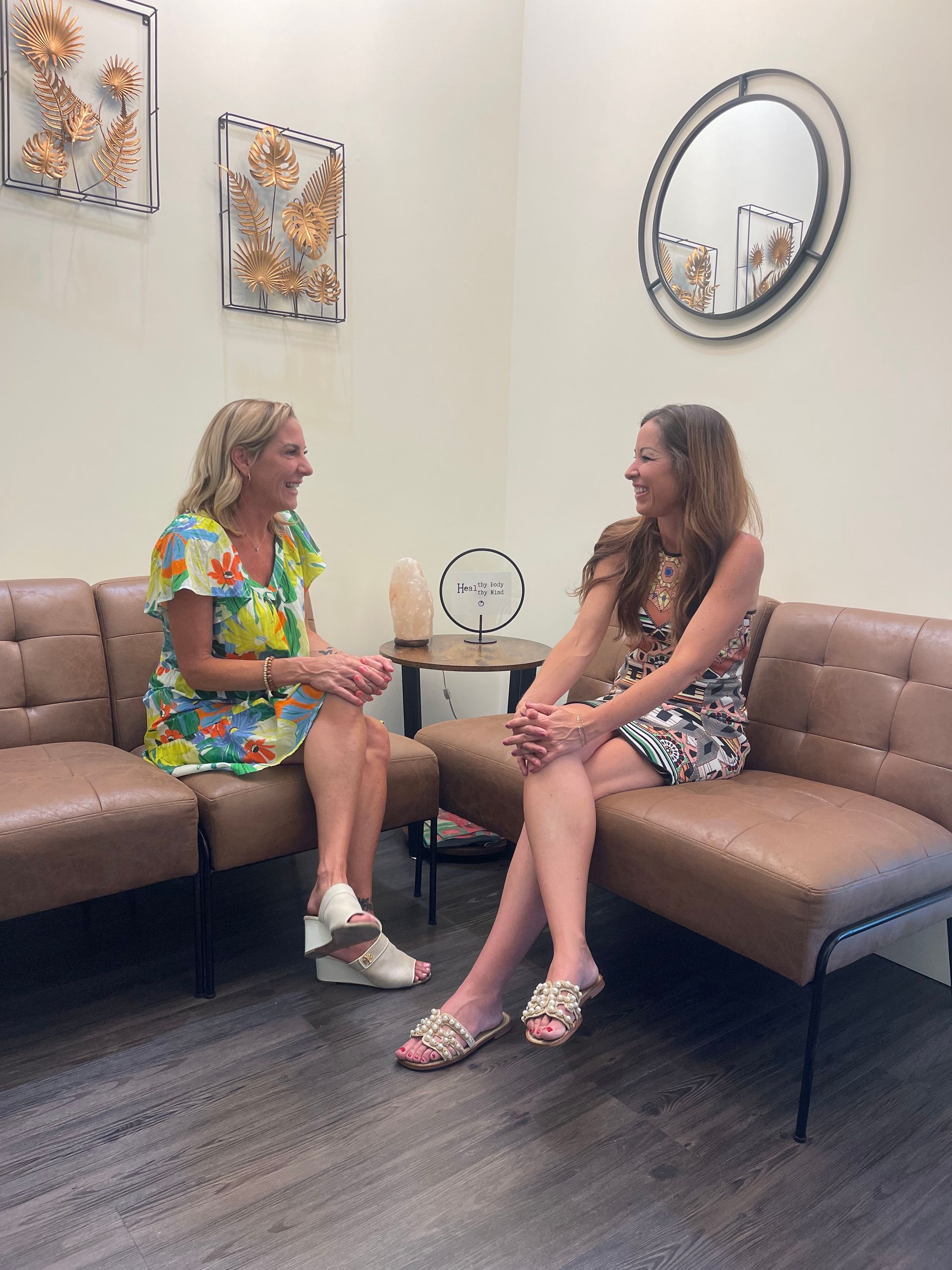 Two women talking on a beige sectional in a bright waiting room with wall art and a round mirror.