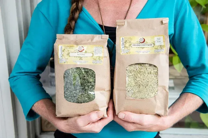 Person holding two brown paper bags of dried herbs, one green and one pale yellow.