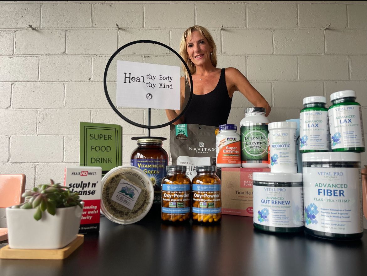 Woman behind a table of supplement bottles and containers, smiling in a bright room with a sign and plants.