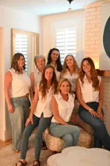 Six women in white tops and jeans pose together in a bright room near a window.