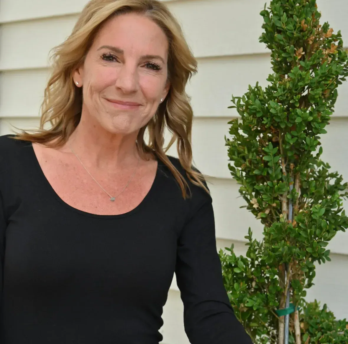 Woman in a black top smiling outdoors beside a trimmed evergreen shrub