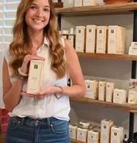 Smiling woman holding a boxed product in a shop with shelves of packaged items behind her