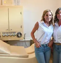Two women standing beside a cream-colored medical examination chair in a clinical room