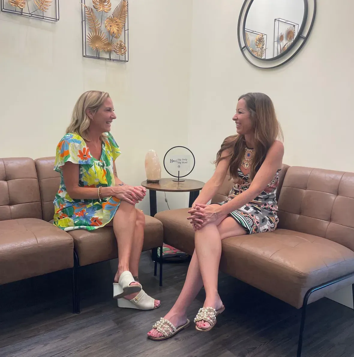 Two women chatting on a tan sectional in a bright waiting room with wall art and a round mirror.