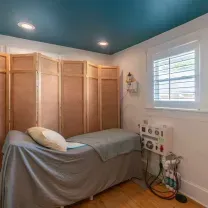 Small room with a massage table, folding privacy screens, white walls, teal ceiling, and a window