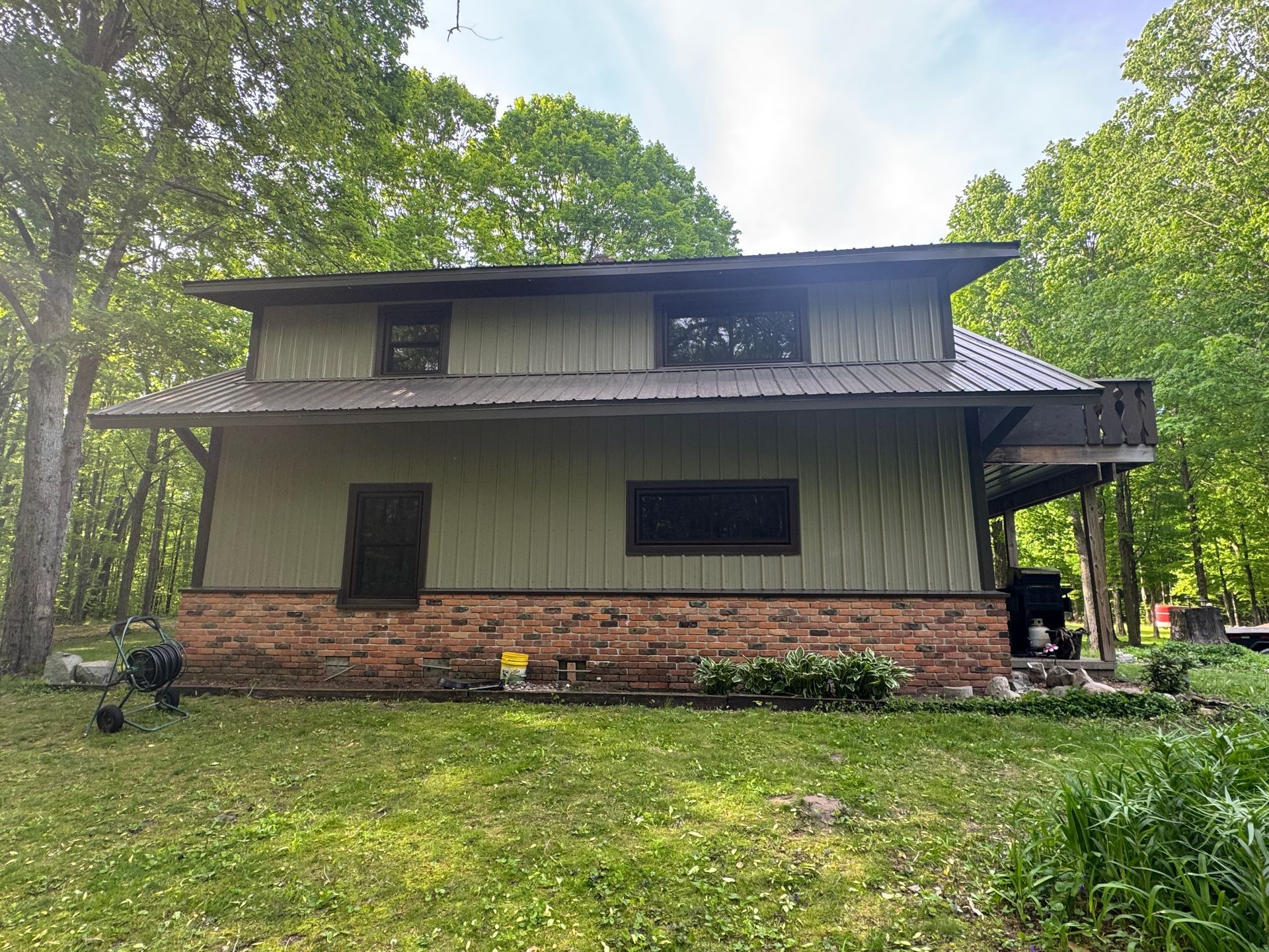Green two-story cabin with brick base, dark roof, and porch in a wooded yard
